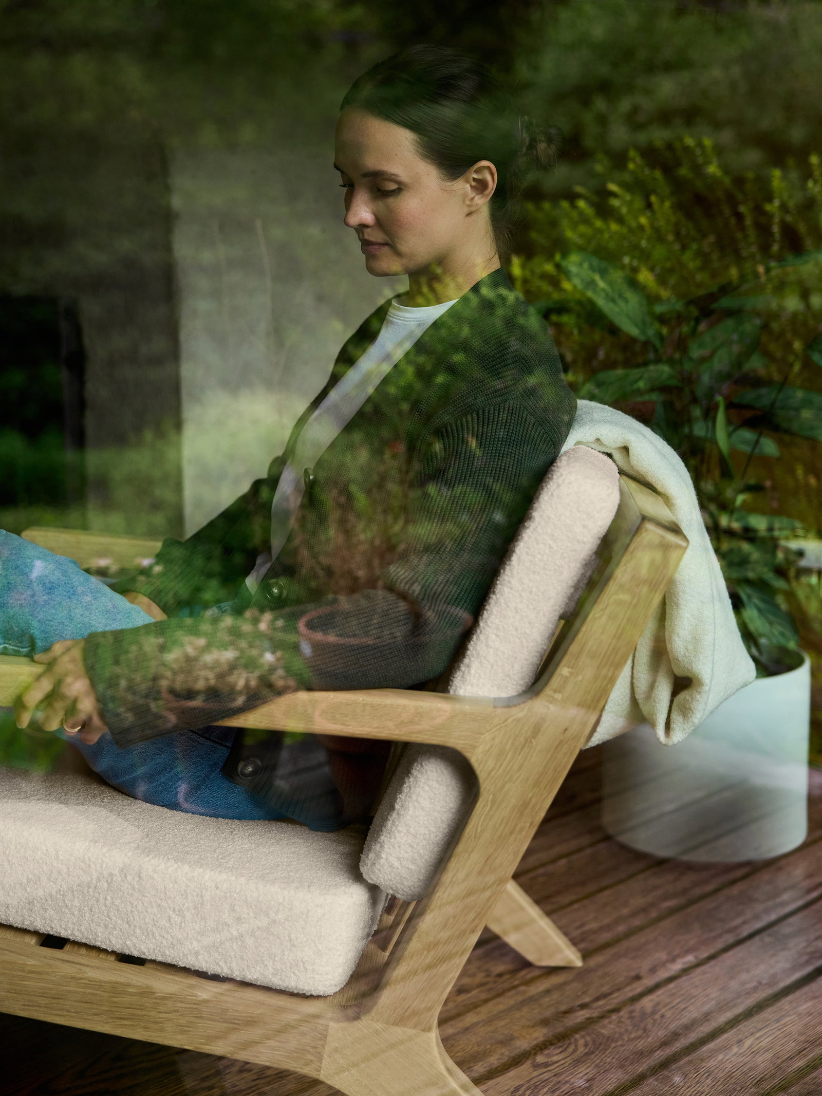 A person sits in a Coronado Lounge Chair by Cozy Earth, featuring a white cushion, while holding a tablet. They are dressed in a dark sweater and light jeans, with a white blanket draped over the chair. Green foliage is visible through the glass window. 