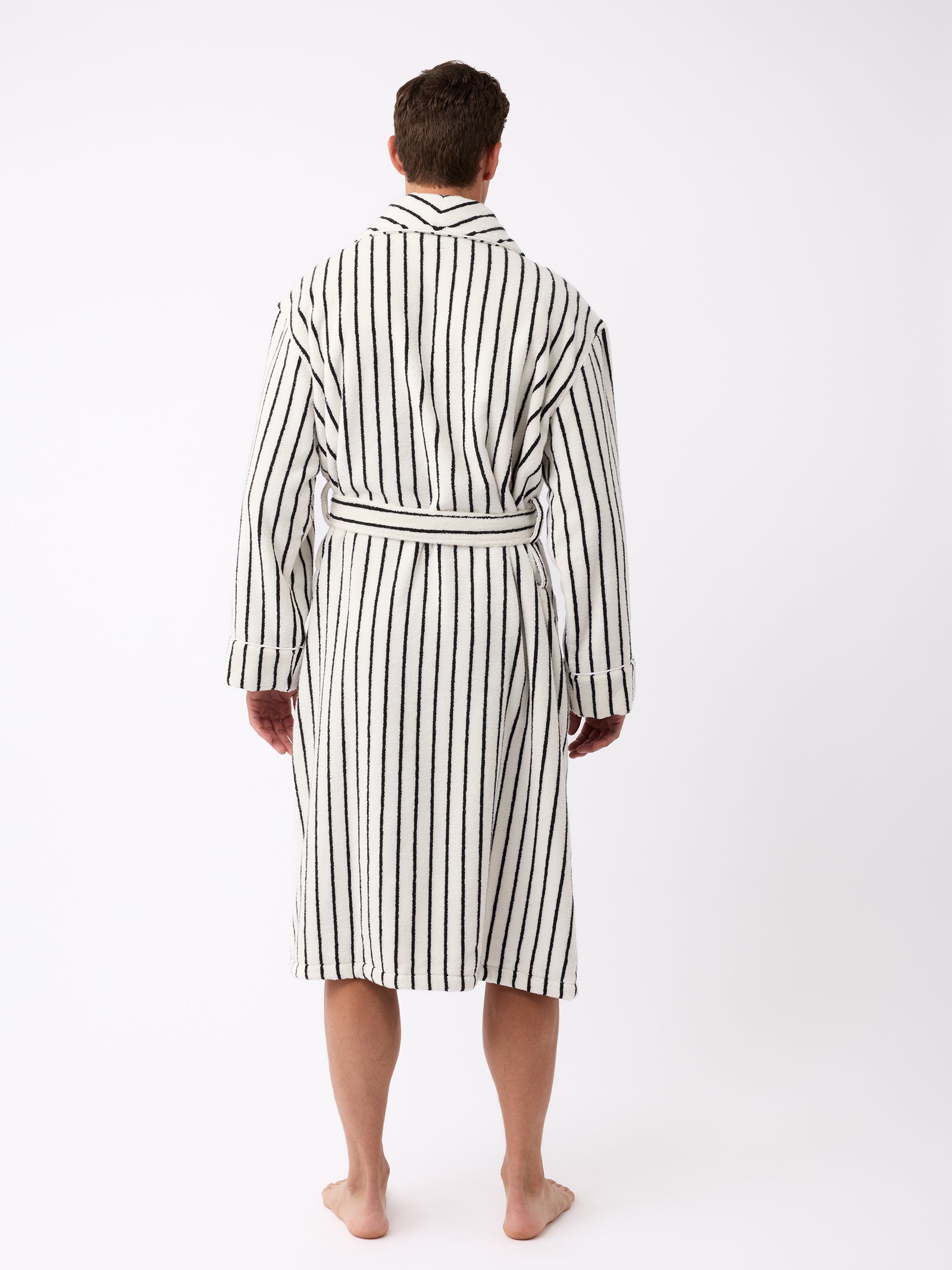 A person is standing barefoot and facing away, wearing the Luxe Bath Robe from Cozy Earth, featuring vertical black stripes. The robe includes a belt tied at the waist, set against a plain white background. 