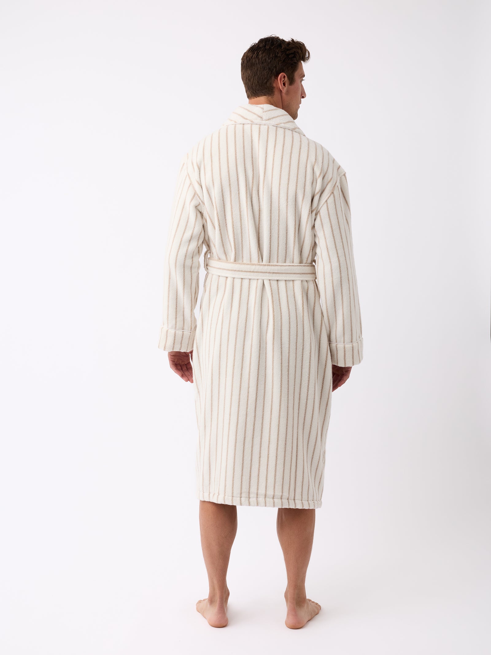 A person is standing barefoot on a white background, facing away and wearing the Luxe Bath Robe by Cozy Earth. The robe features long vertical stripes and a belt tied at the waist, all set in a minimal and well-lit environment. 