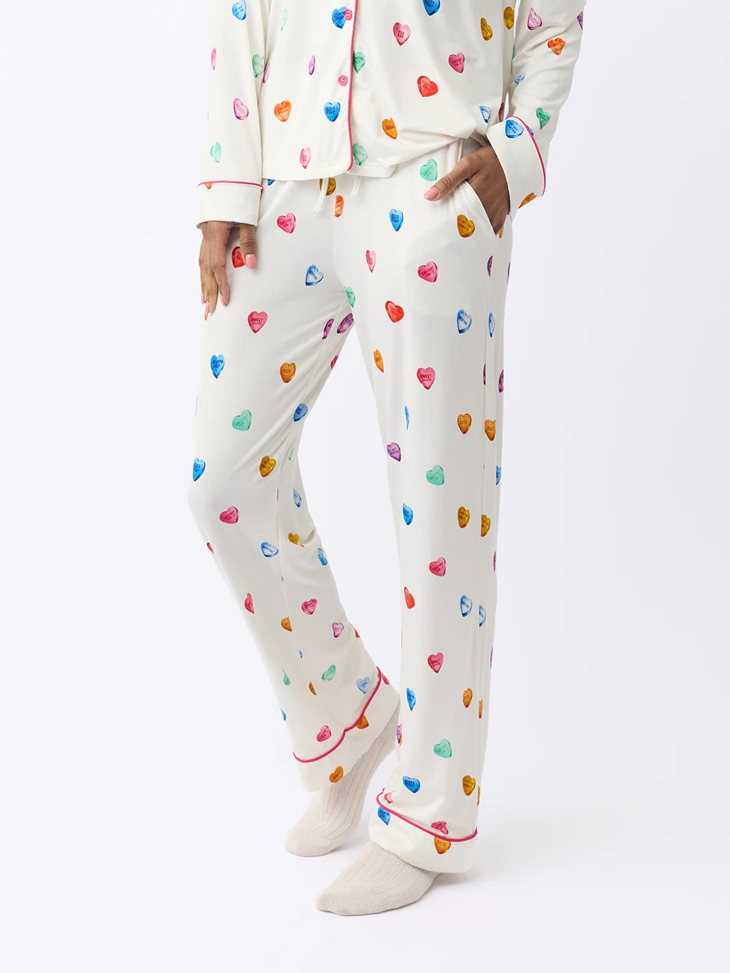 A person models Cozy Earth Women's Stretch-Knit Bamboo Pajama Pant with a colorful heart pattern, red trim, and cream-colored socks against a plain white background. |Color:Sleepy Hearts