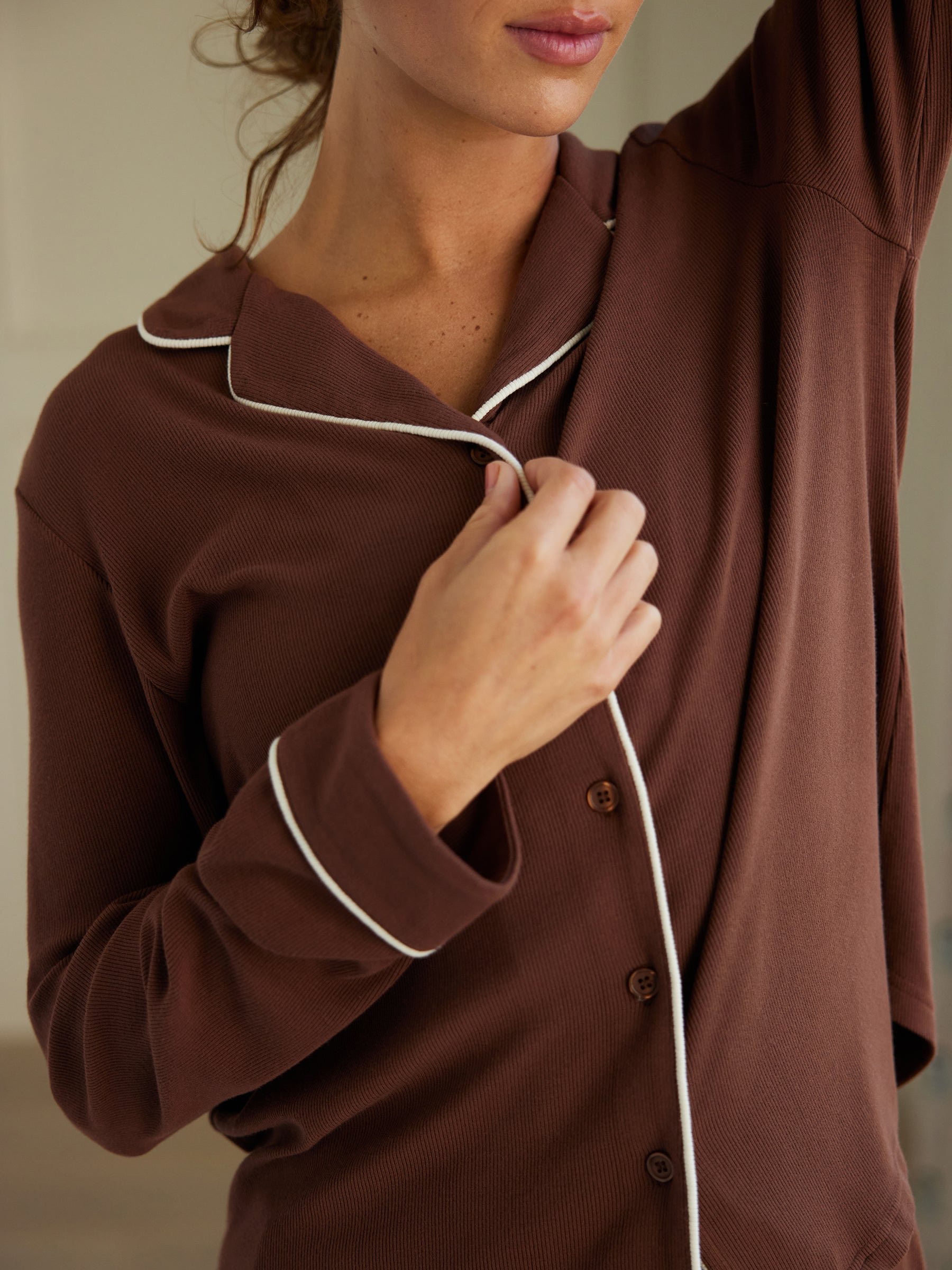 A woman wearing the Cozy Earth Women's Bamboo Rib Knit Classic Long Sleeve Pajama Top in brown with white piping holds her collar while raising her other arm. The image highlights the chest and arm area, with the person's face partially visible. The background is blurred, drawing attention to her attire. 