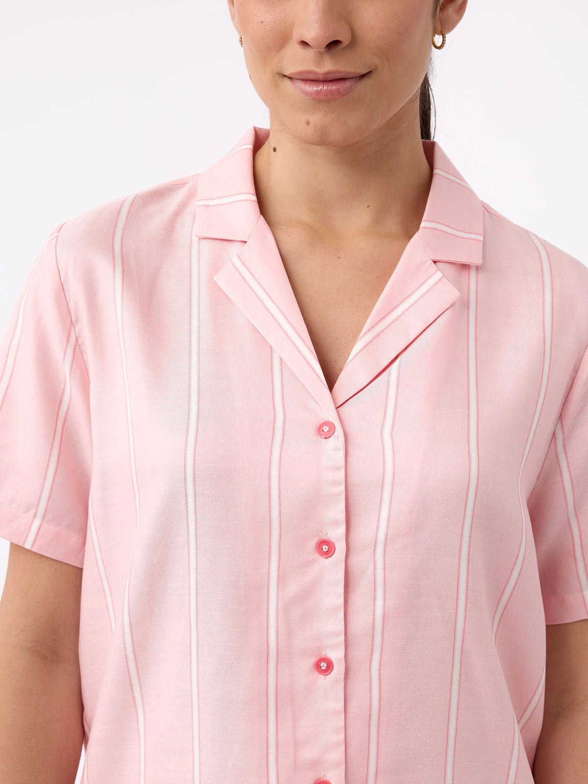 A person wearing a Cozy Earth Women's Soft Woven Short Sleeve Pajama Set in pink with thin white stripes is visible from shoulders to mid-torso. The shirt features a notched collar, and the individual has long hair tied back, standing against a plain white background. 