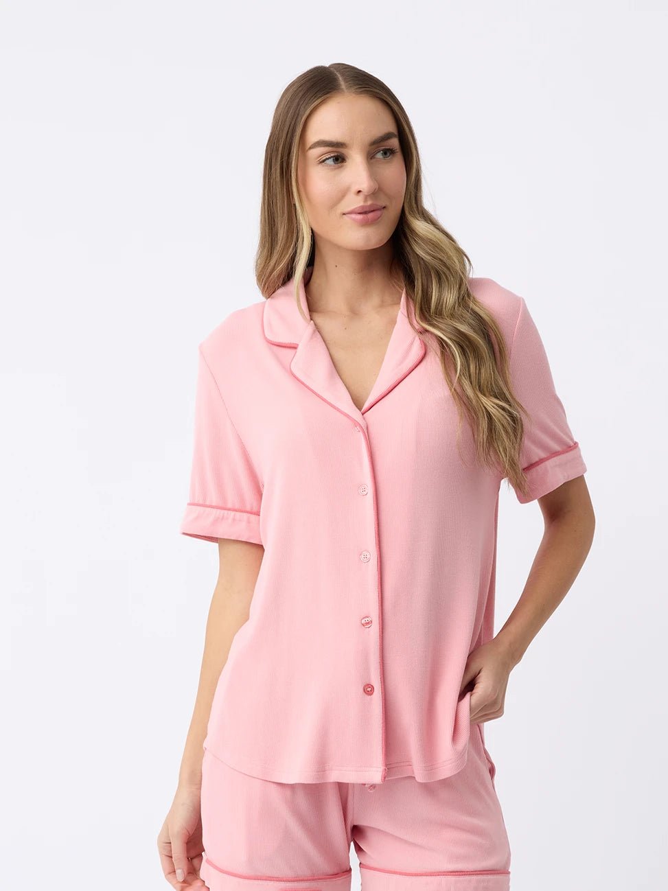 A woman with long hair against a white background wears the Women's Bamboo Rib-Knit Classic Short Sleeve Pajama Top in pink by Cozy Earth, paired with shorts. She has a relaxed expression and her right hand is near her waist. 