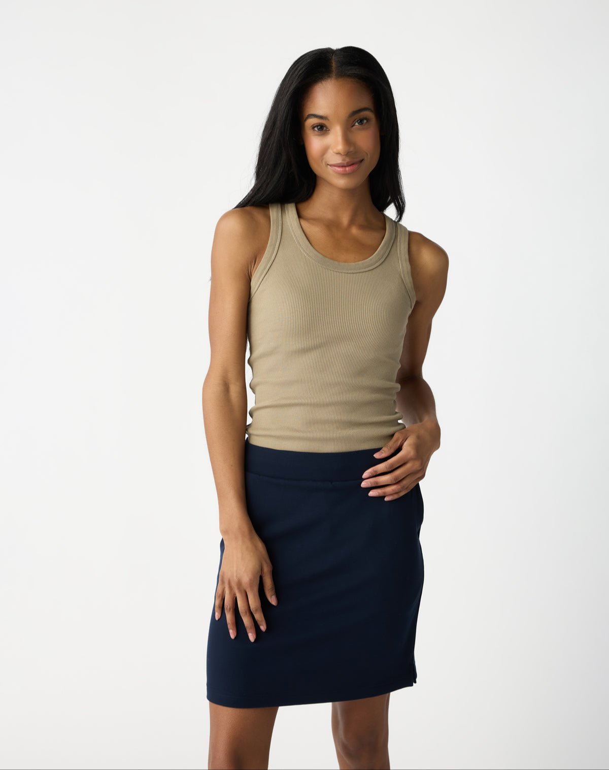 A person with long dark hair strikes a confident pose against a light grey backdrop, wearing a beige tank top and Cozy Earth's Women's Brushed Bamboo Skirt in dark blue. With a slight smile, hand on hip, they exude a relaxed vibe. 