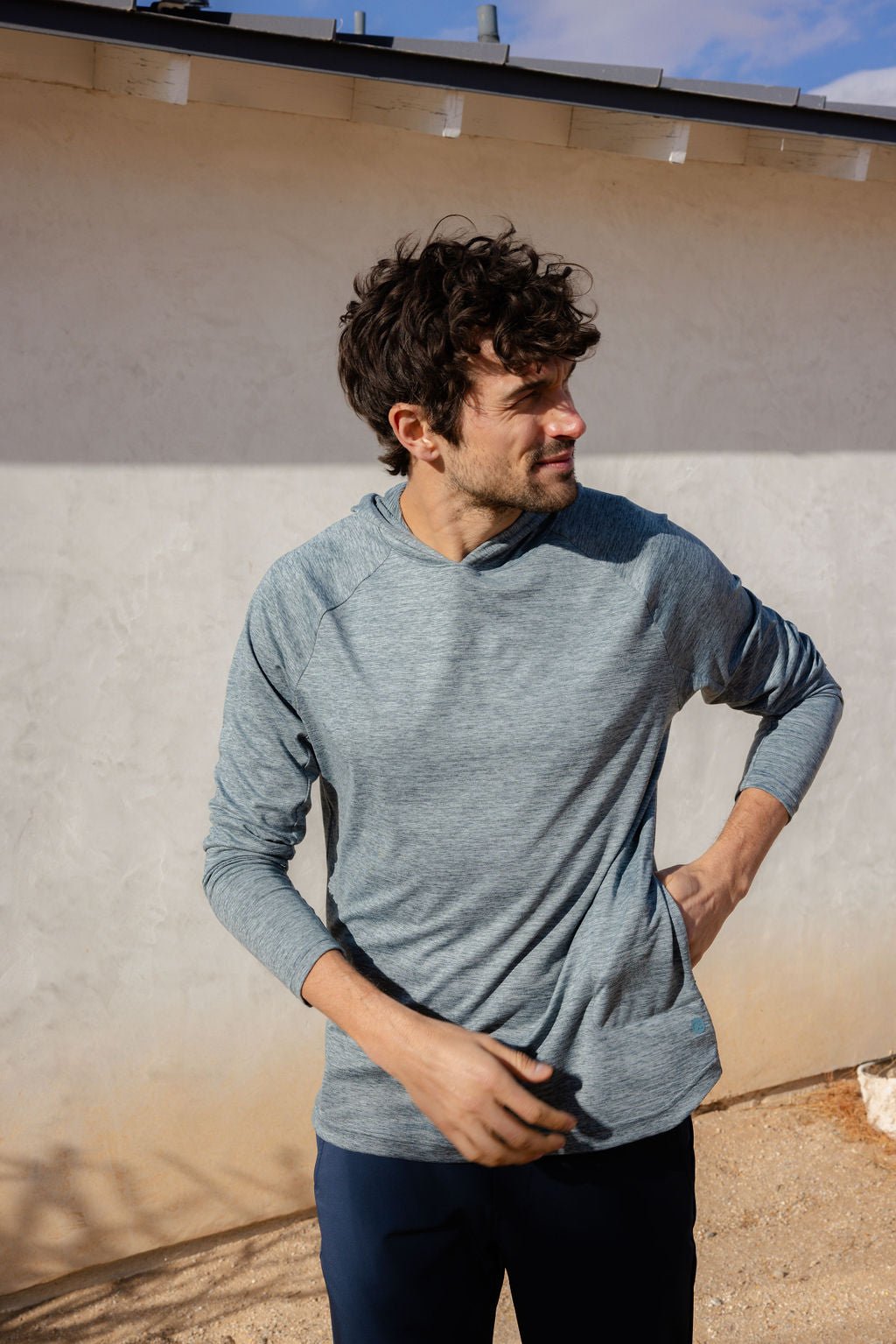 A man with curly hair stands outside, wearing a Cozy Earth Men's Lightweight Performance Hoodie in gray and dark pants. He looks to his left, one hand on his hip, and the other by his side. A shadow is cast on the white wall behind him. 
