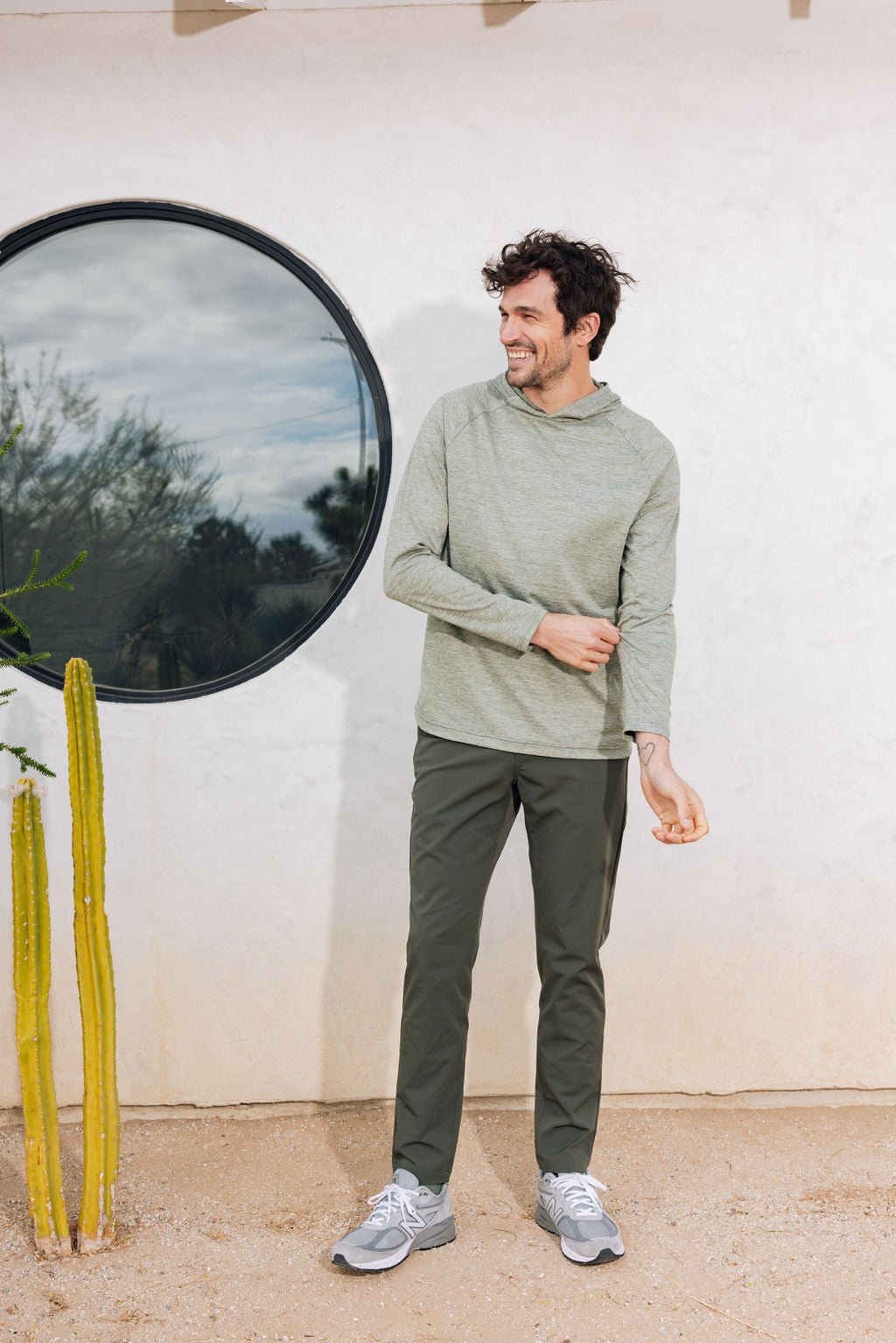 A man in a Cozy Earth Men's Lightweight Performance Hoodie and dark pants stands outside, smiling near a circular window. He's adjusting his sleeve while wearing gray sneakers, with a tall cactus beside him against a light-colored wall. 