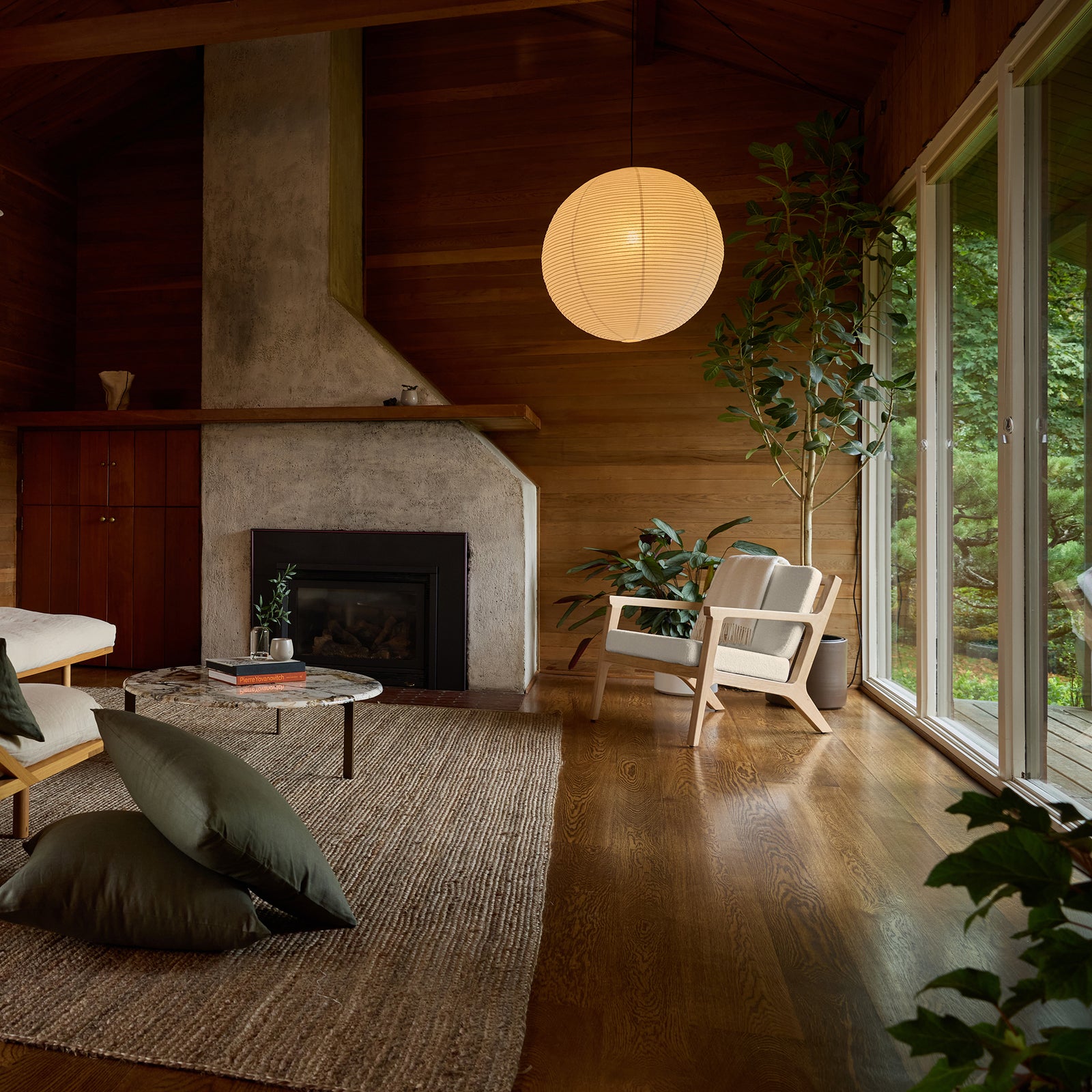 A cozy living room with a modern fireplace, wooden walls, and a large round paper lantern hanging from the ceiling. It features the Coronado Lounge Chair by Cozy Earth, a round coffee table, green cushions, plants, and a large window showcasing a lush outdoor scene. 