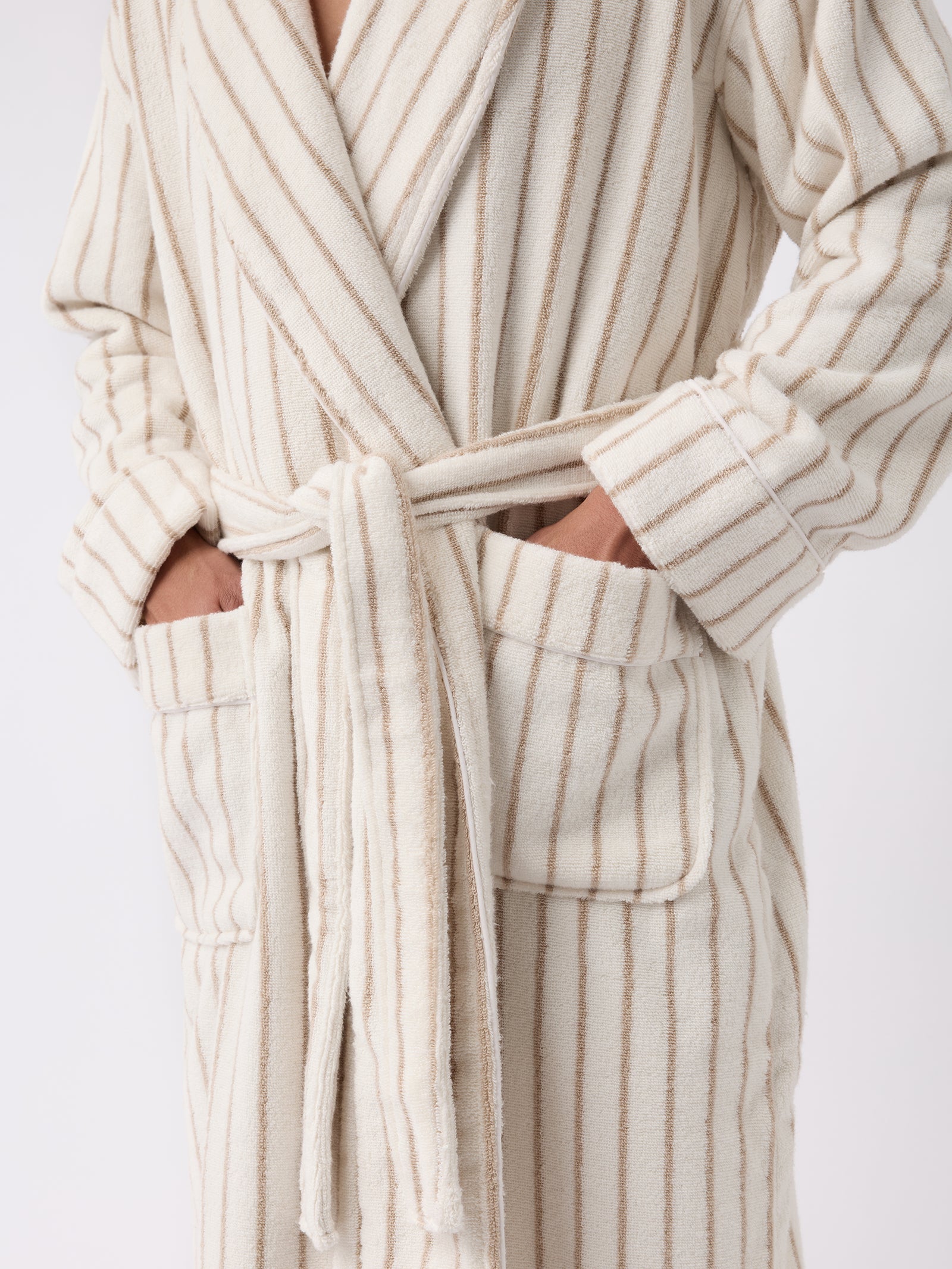 A person wearing the Luxe Bath Robe by Cozy Earth, which showcases a beige and white striped design with hands in its pockets. The robe includes a belt tied at the waist against a plain light-colored background. 