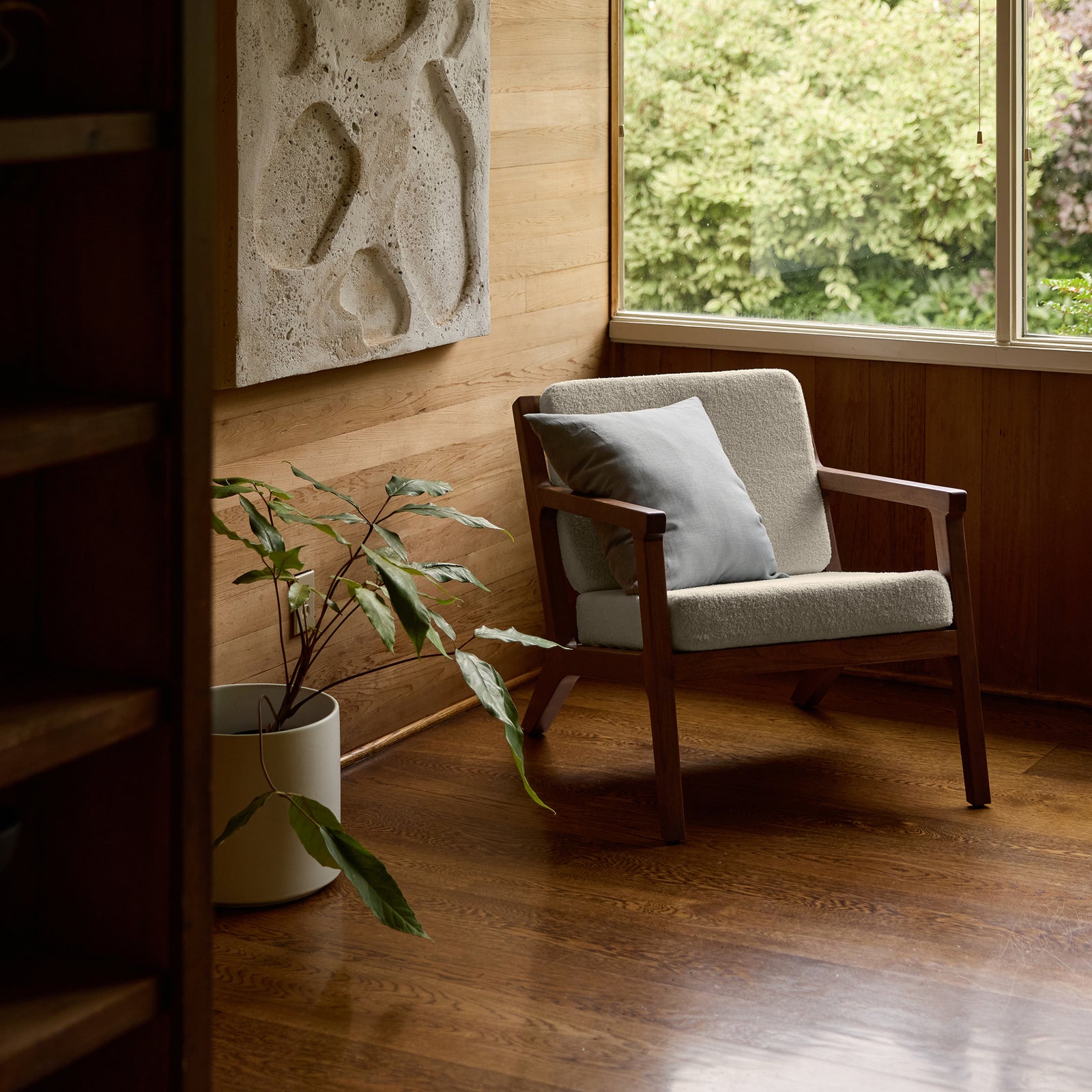 In a cozy room corner, the Coronado Lounge Chair by Cozy Earth, featuring a gray cushion, is placed on a polished wooden floor. Nearby, a tall plant in a white pot and textured wall art complement the setting as sunlight streams through a window showcasing greenery outside. 