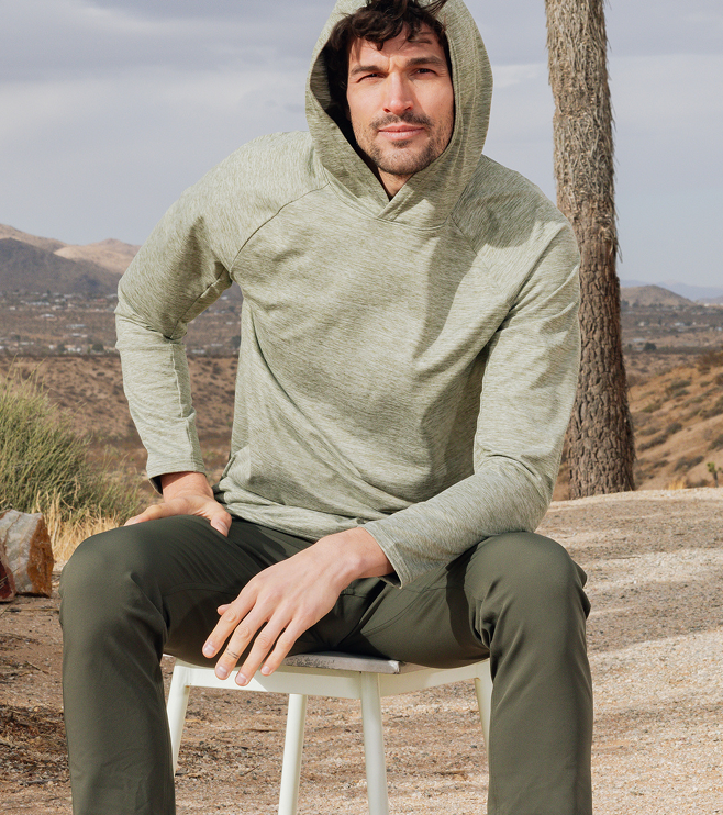 Man sitting on a white chair outdoors wearing an olive green hoodie and dark green pants. Desert landscape with mountains and trees in the background. Cloudy sky overhead.
