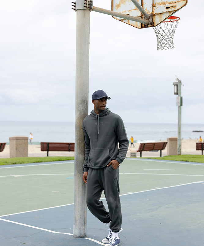 A person in a dark hoodie and sweatpants leans against a basketball hoop pole on an outdoor court by the beach. They wear a cap and sneakers. The sky is cloudy, and the ocean is visible in the background. Benches and a person walking can be seen.