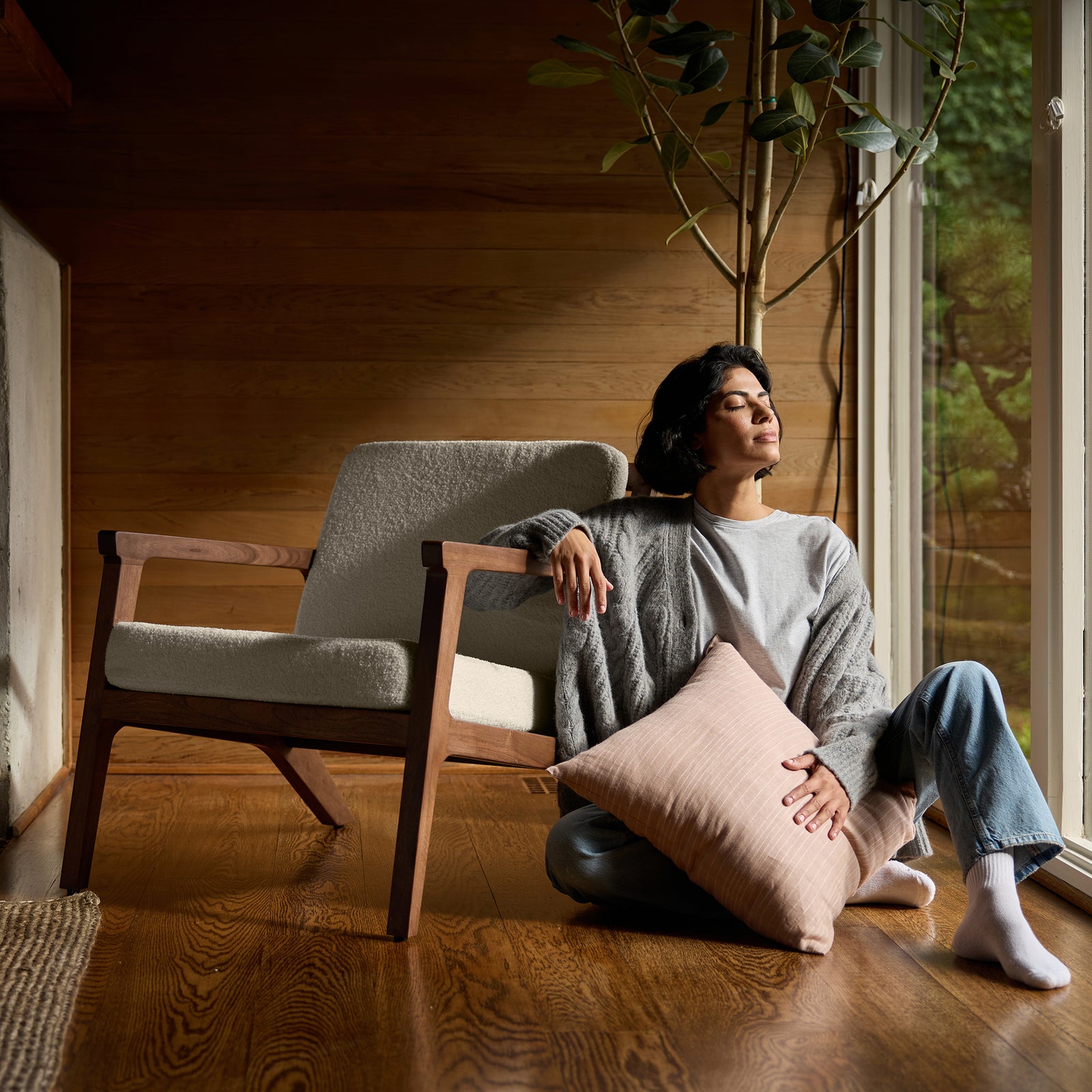 A person relaxes by a large window, sitting on the floor and leaning against a Coronado Lounge Chair by Cozy Earth, which features a soft cushion. They are holding a pink pillow and looking serene as sunlight filters through the window, highlighting the cozy wooden interior. 