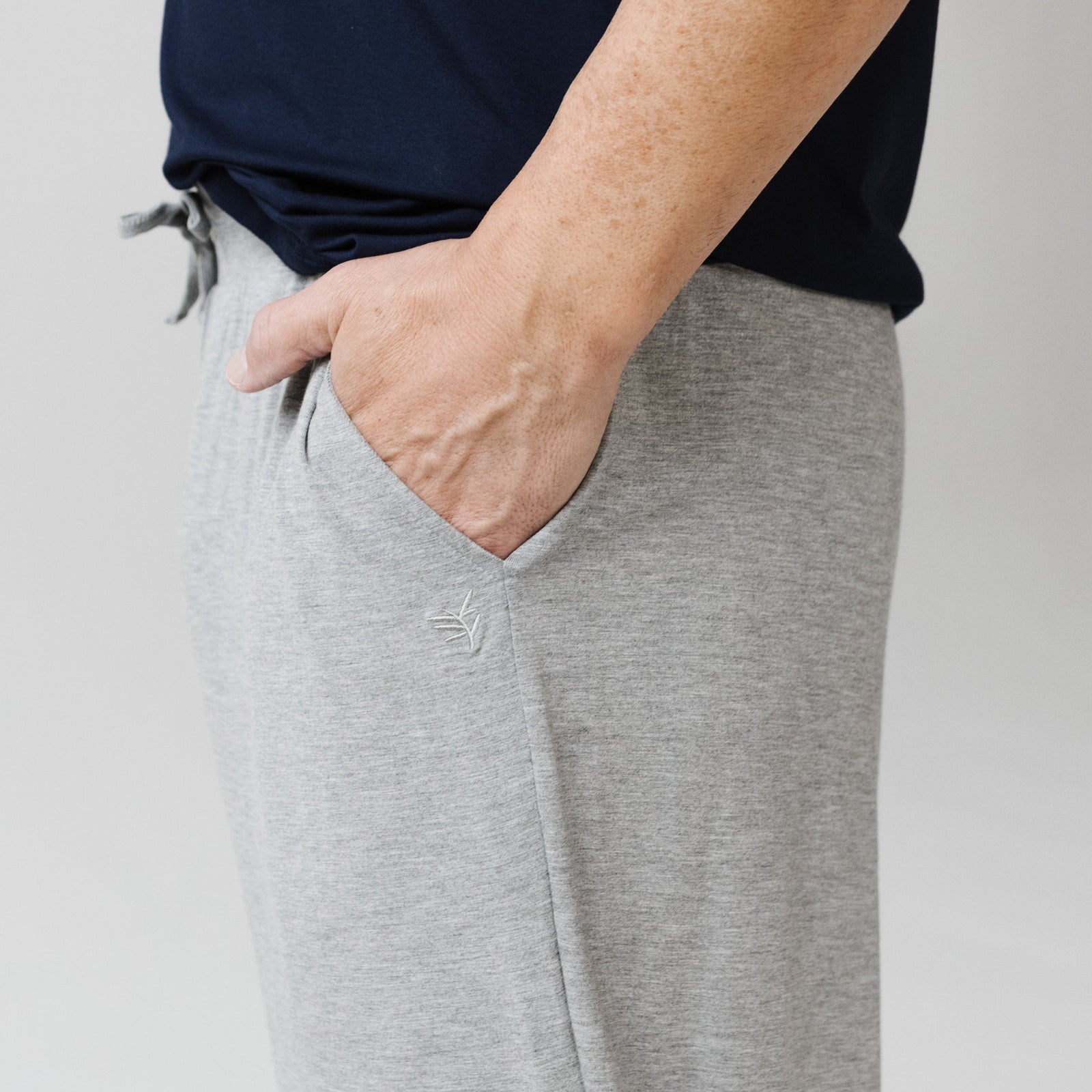 A person stands with one hand in the pocket of their light gray pajama pants. They are wearing the men's stretch-knit long sleeve navy blue shirt from Cozy Earth. The background is plain and neutral, emphasizing the clothing. 