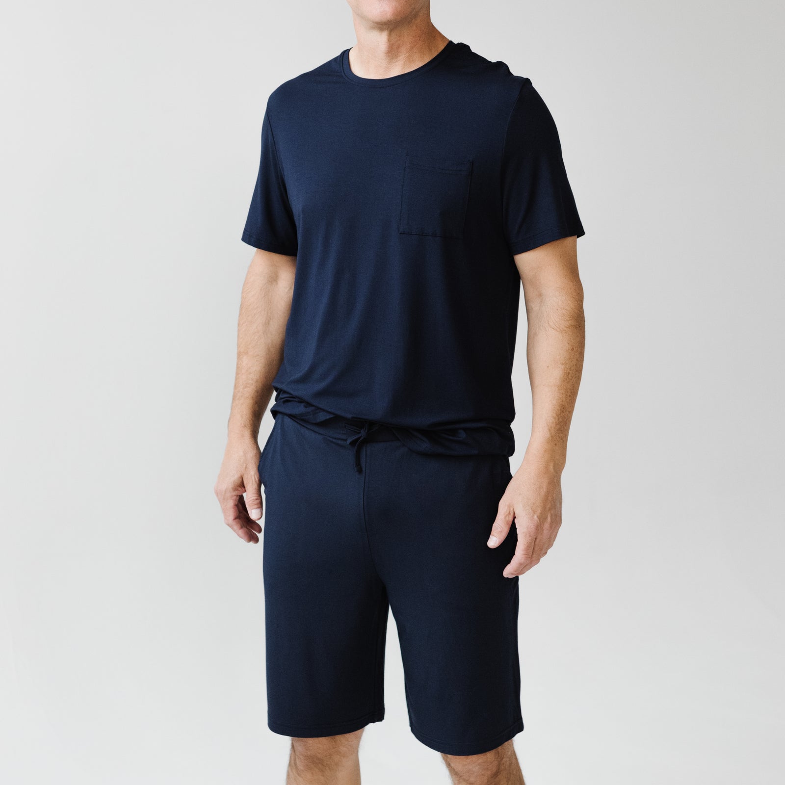 A person is wearing the Men's Stretch-Knit Short Sleeve Bamboo Pajama Set from Cozy Earth, featuring a dark blue top with a pocket on the left side and matching shorts. The outfit offers a casual look against a plain, light-colored background.