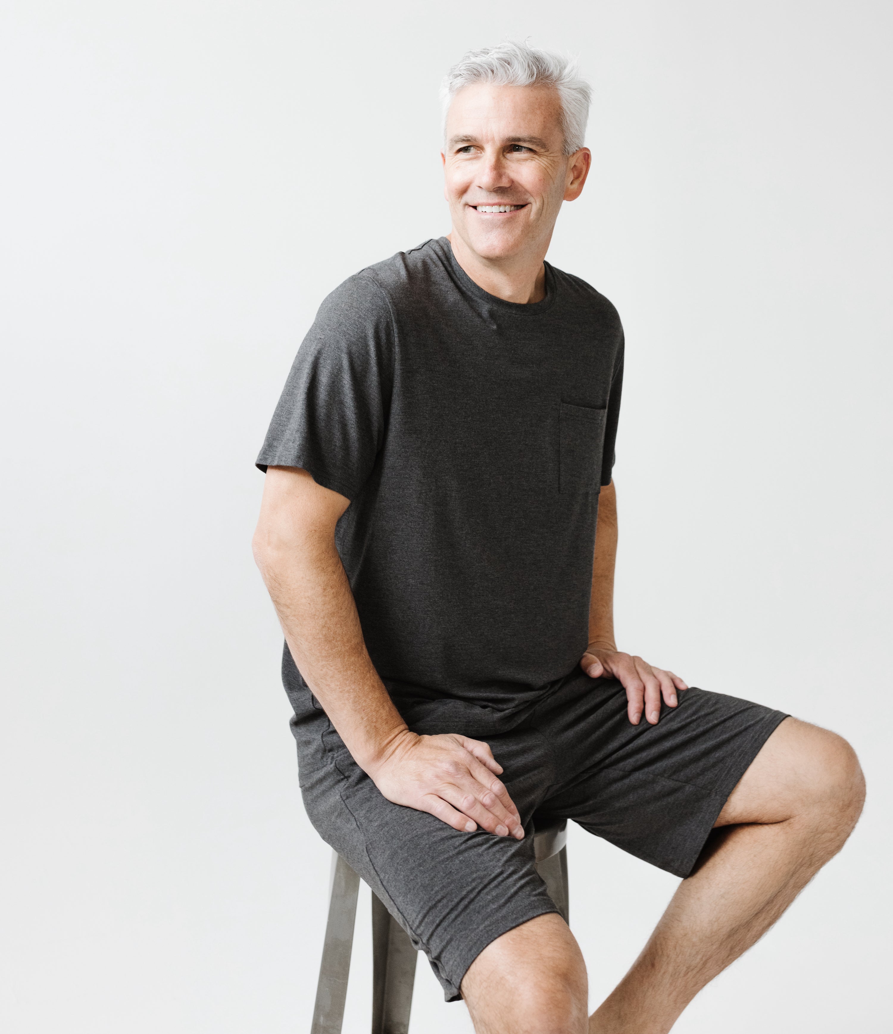 A smiling man with gray hair sits on a stool, comfortably dressed in the Cozy Earth Men's Stretch-Knit Short Sleeve Bamboo Pajama Set. The plain white background enhances the clean and minimal appearance of the scene.