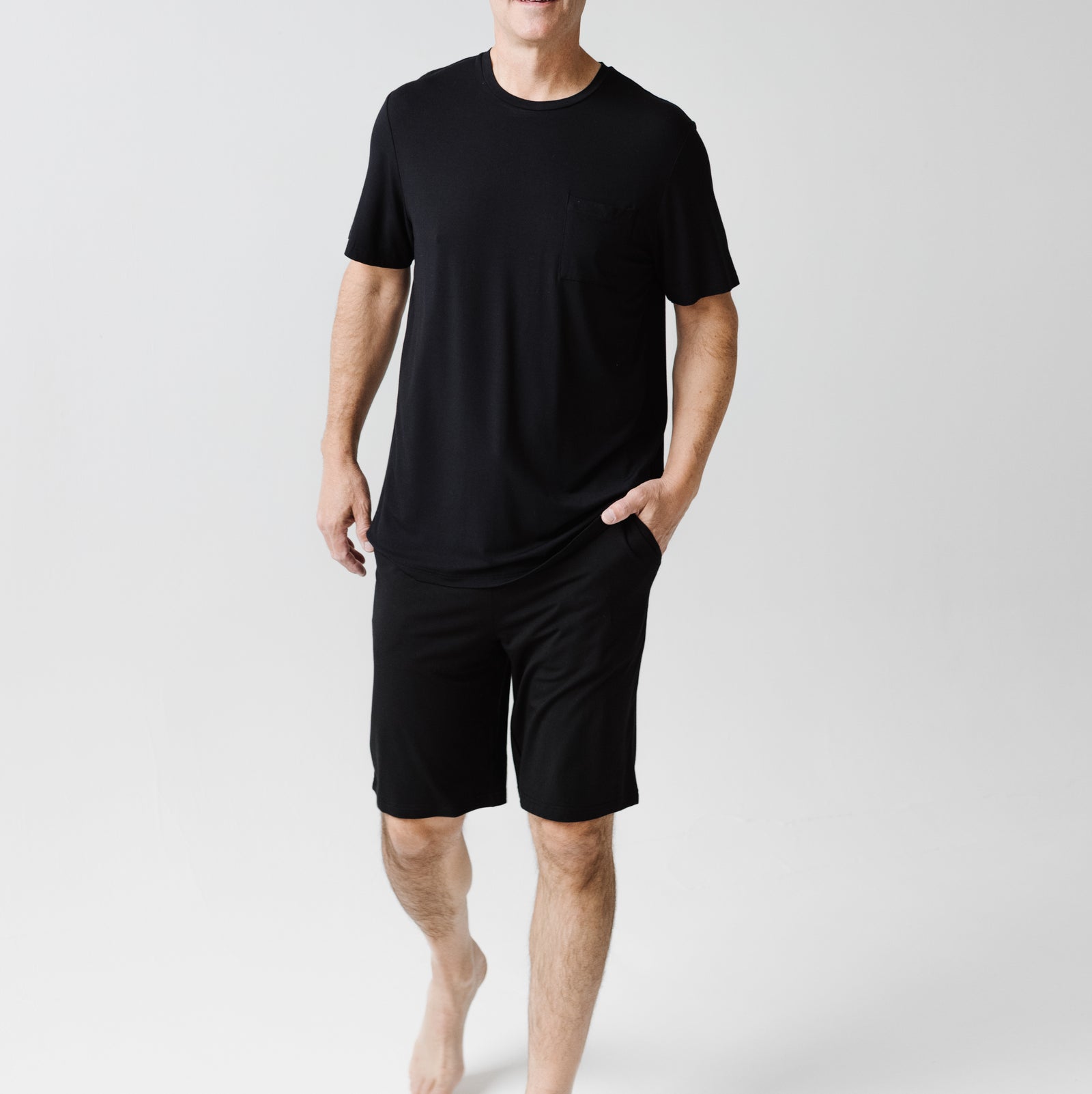 A person wearing a Men's Stretch-Knit Short Sleeve Bamboo Pajama Set from Cozy Earth walks barefoot on a plain white background. Their left hand rests in their pocket, and they appear relaxed and casual.