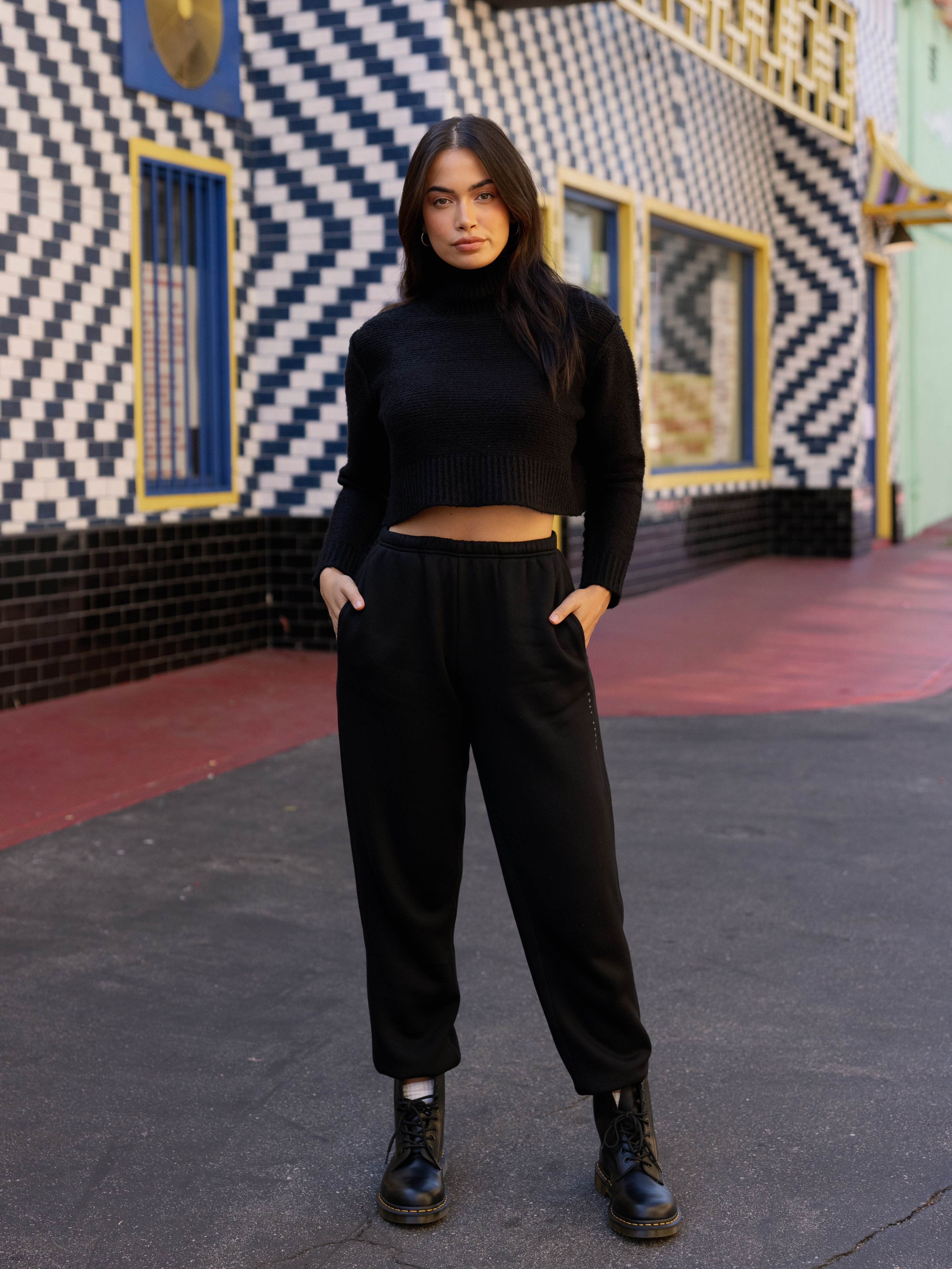 Black CityScape Joggers. The Joggers are being worn by a female model. The photo is taken with the models hands by the pocket of the joggers. The back ground is a downtown area. 