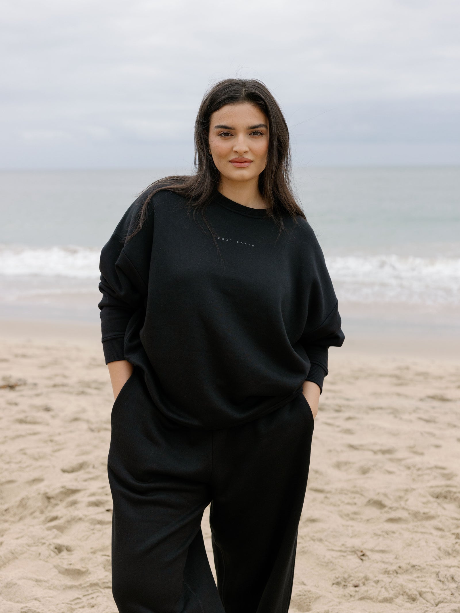 Black CityScape Pullover Crew. The Pullover is being worn by a female model. Accompanying city scape clothing is being worn to complete the look of the outfit. The photo was taken with a sandy beach background. 