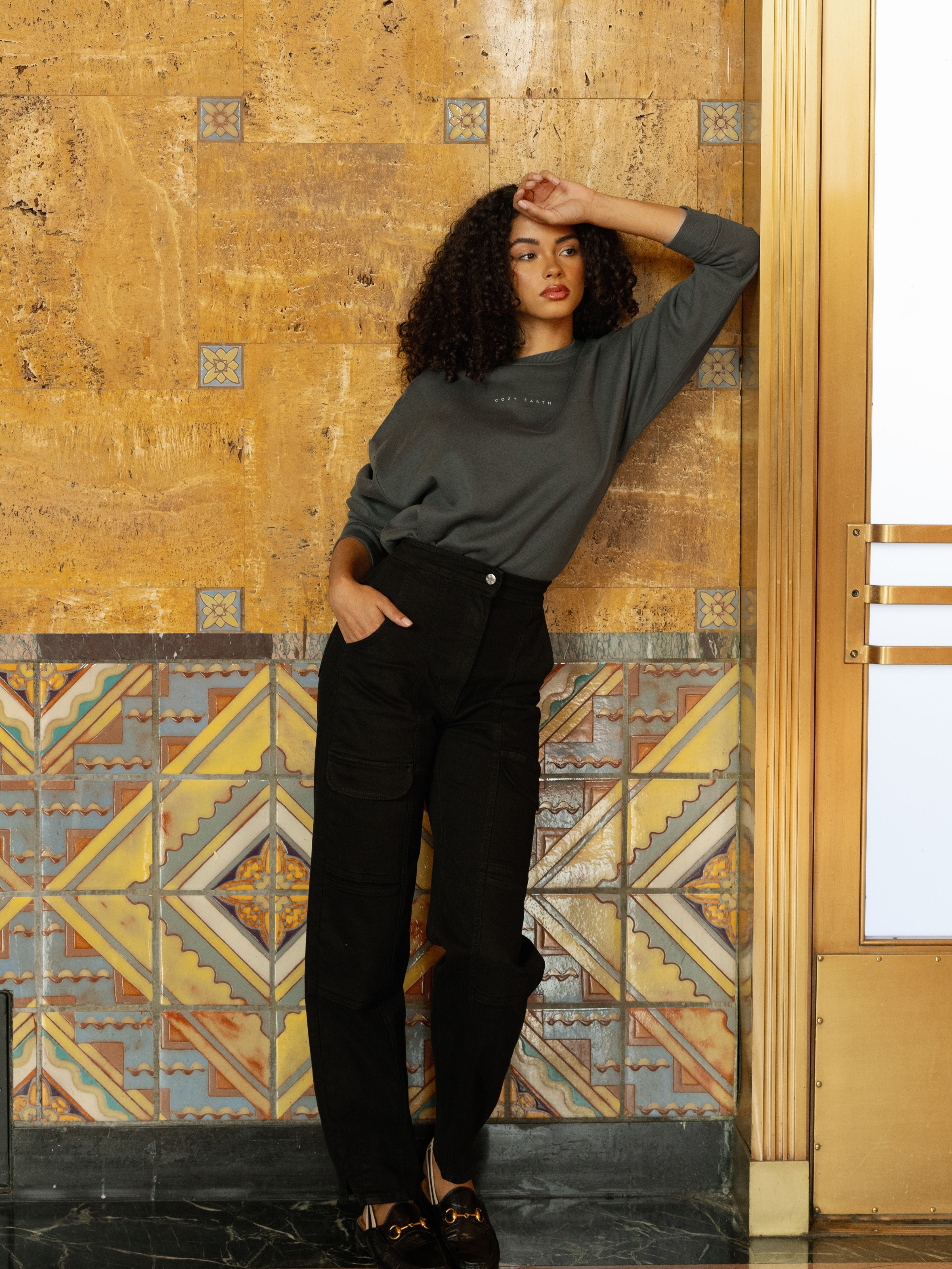Storm CityScape Pullover Crew. The Pullover is being worn by a female model. An accompanying pair of black jeans are being worn to complete the look of the outfit. The photo was taken with a city building background. 
