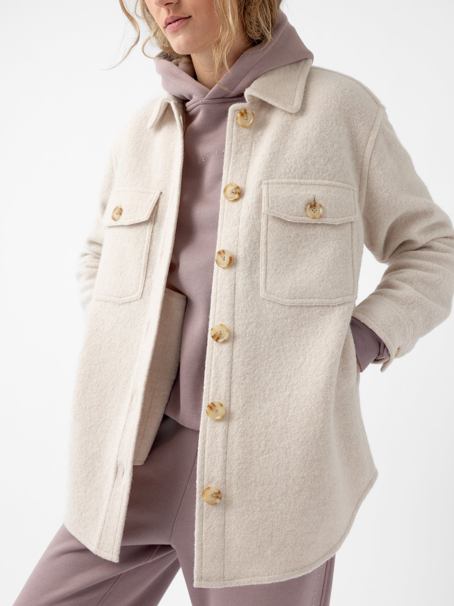 A person wearing a Cozy Earth Women's Boucle Shacket in beige over a mauve hoodie and matching pants. The shacket features large buttons and two front pockets, with their hands tucked into the pockets. The background is plain white.  |Color:Alabaster