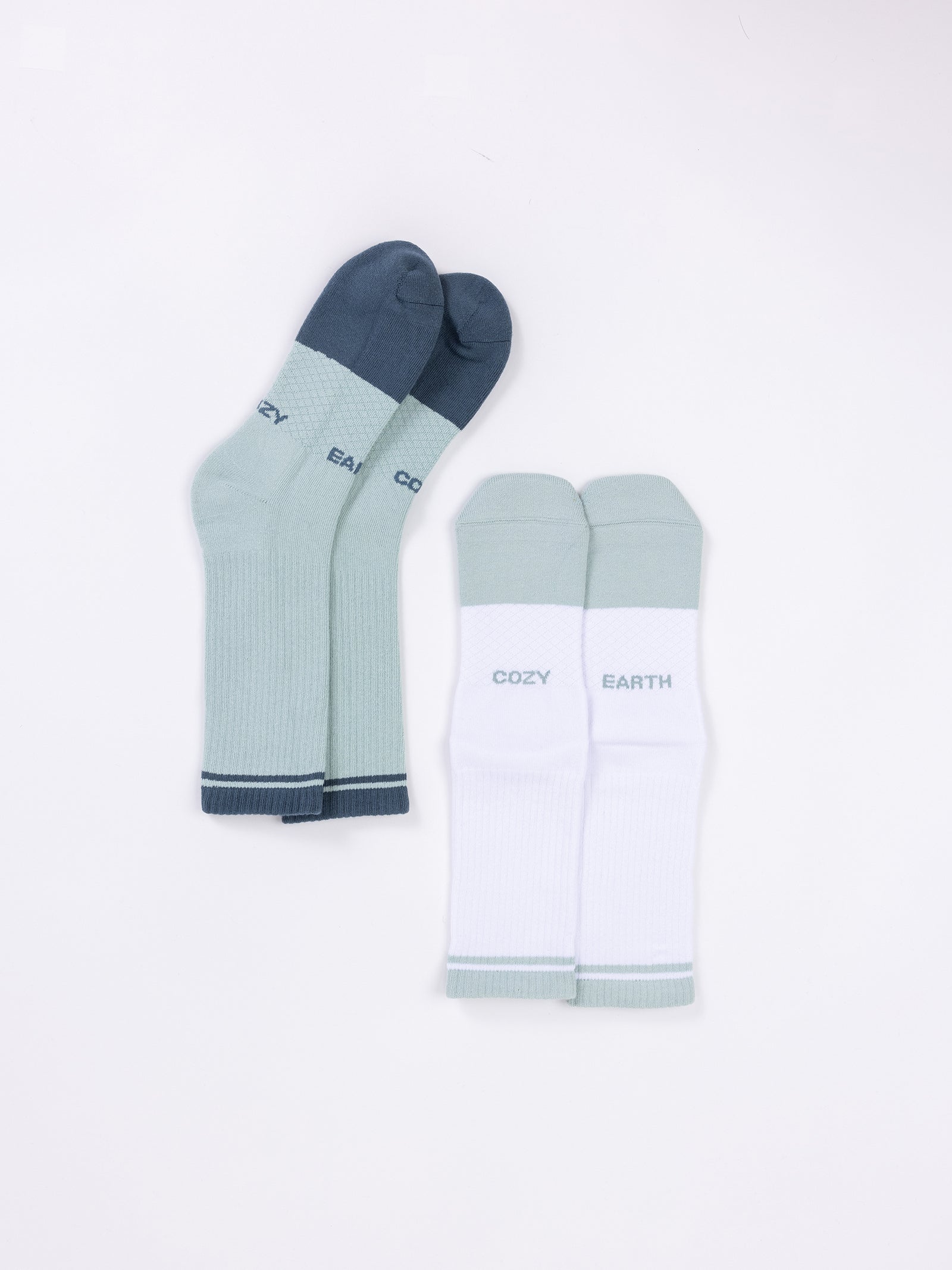 Two pairs of Essential Calf Socks are displayed on a white background. One pair is light green with dark green toes, heels, and cuffs, featuring the word "COZY" near the ankle. The other pair is white with light green toes, heels, and cuffs, featuring the words "COZY EARTH" from Cozy Earth brand. 