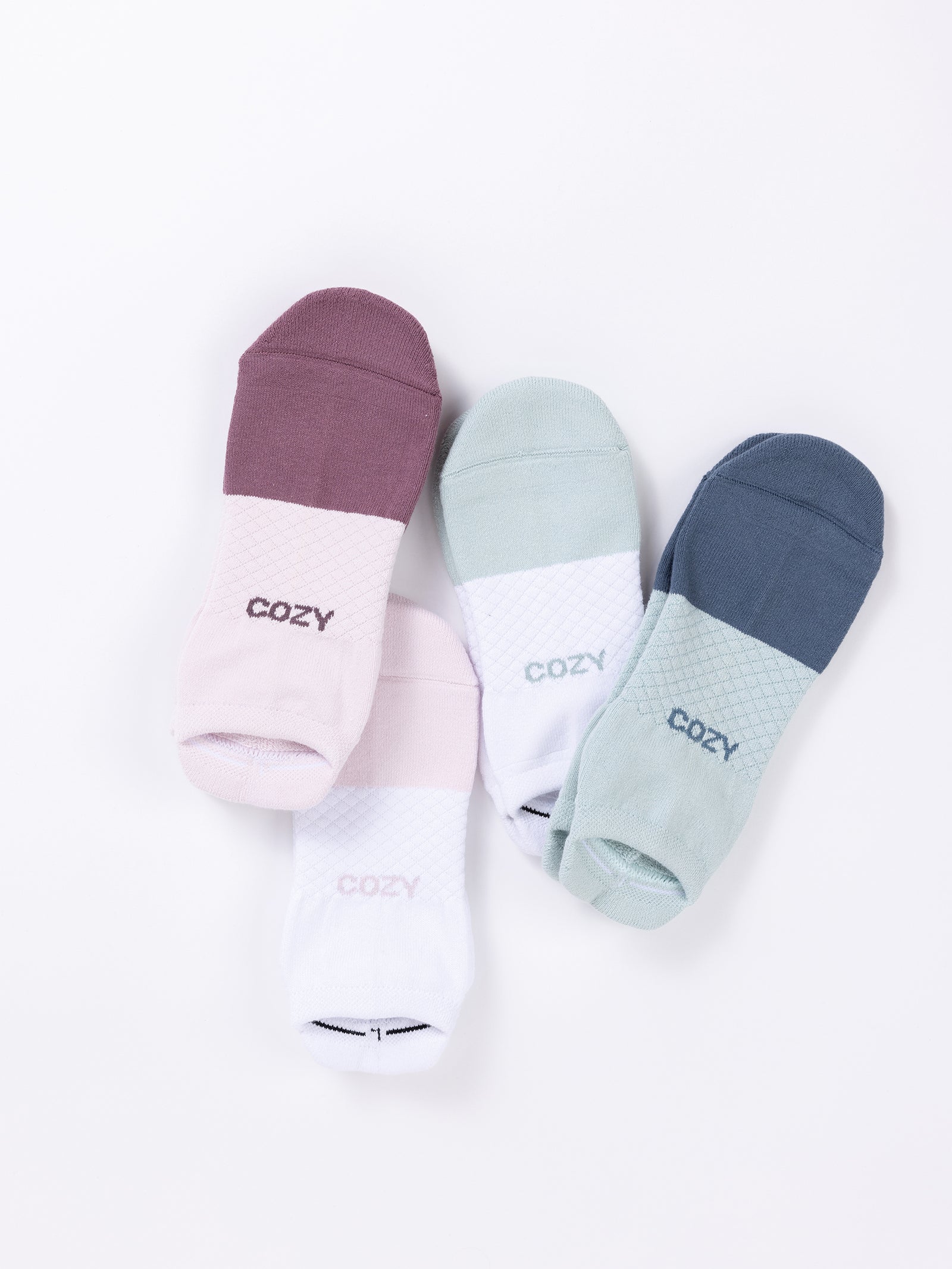 The Essential Ankle Sock 4-Pack from Cozy Earth on a white background. Each of the four pairs features a different color scheme, with one section in a lighter shade and the other in a darker shade. The socks are emblazoned with the word "COZY" in bold letters near the toes. 