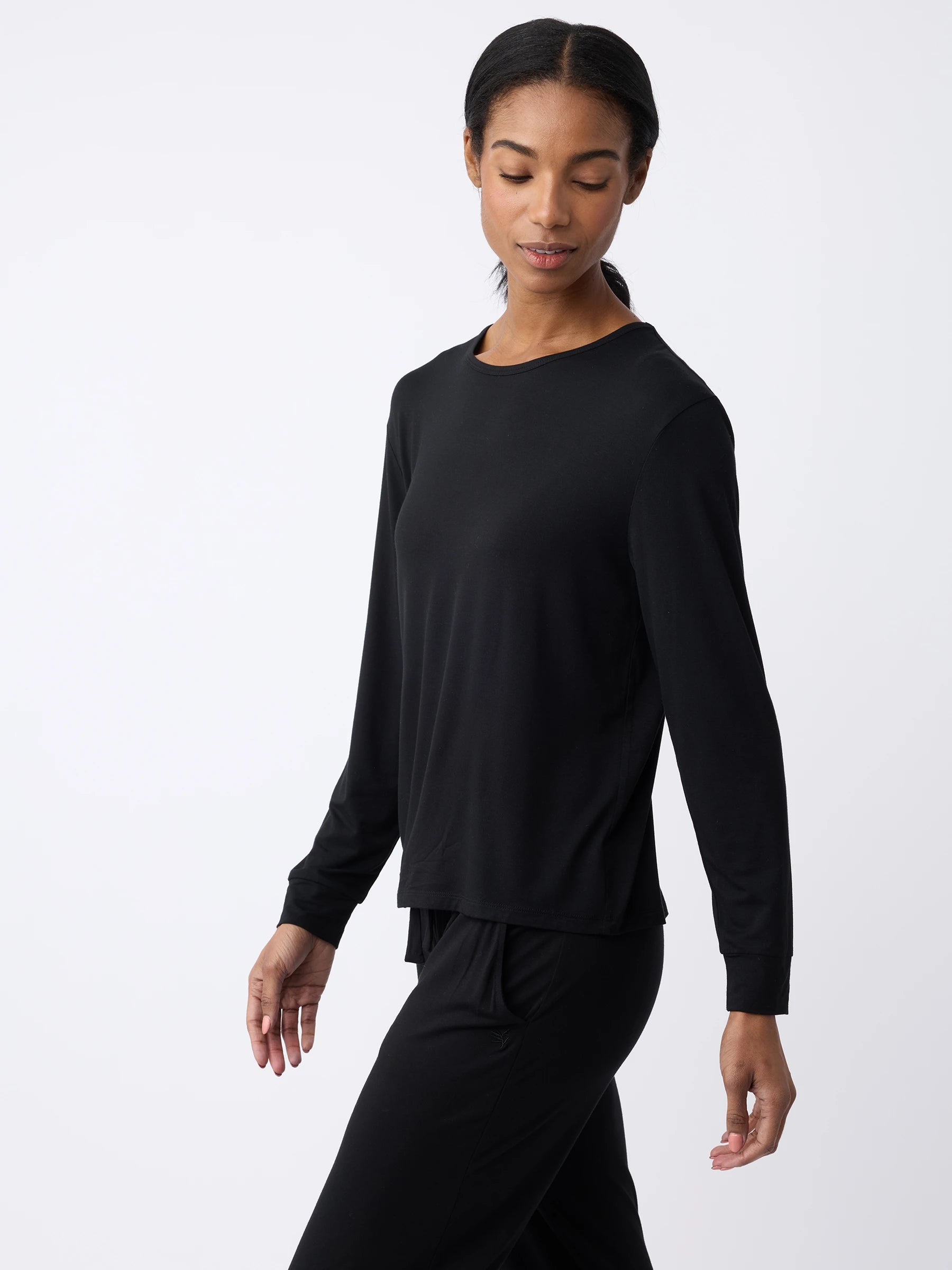 A woman is wearing the Cozy Earth Women's Stretch Knit Bamboo Long Sleeve Lounge Tee and matching pants, walking while looking slightly down. The background is plain and light-colored. |Color:Black