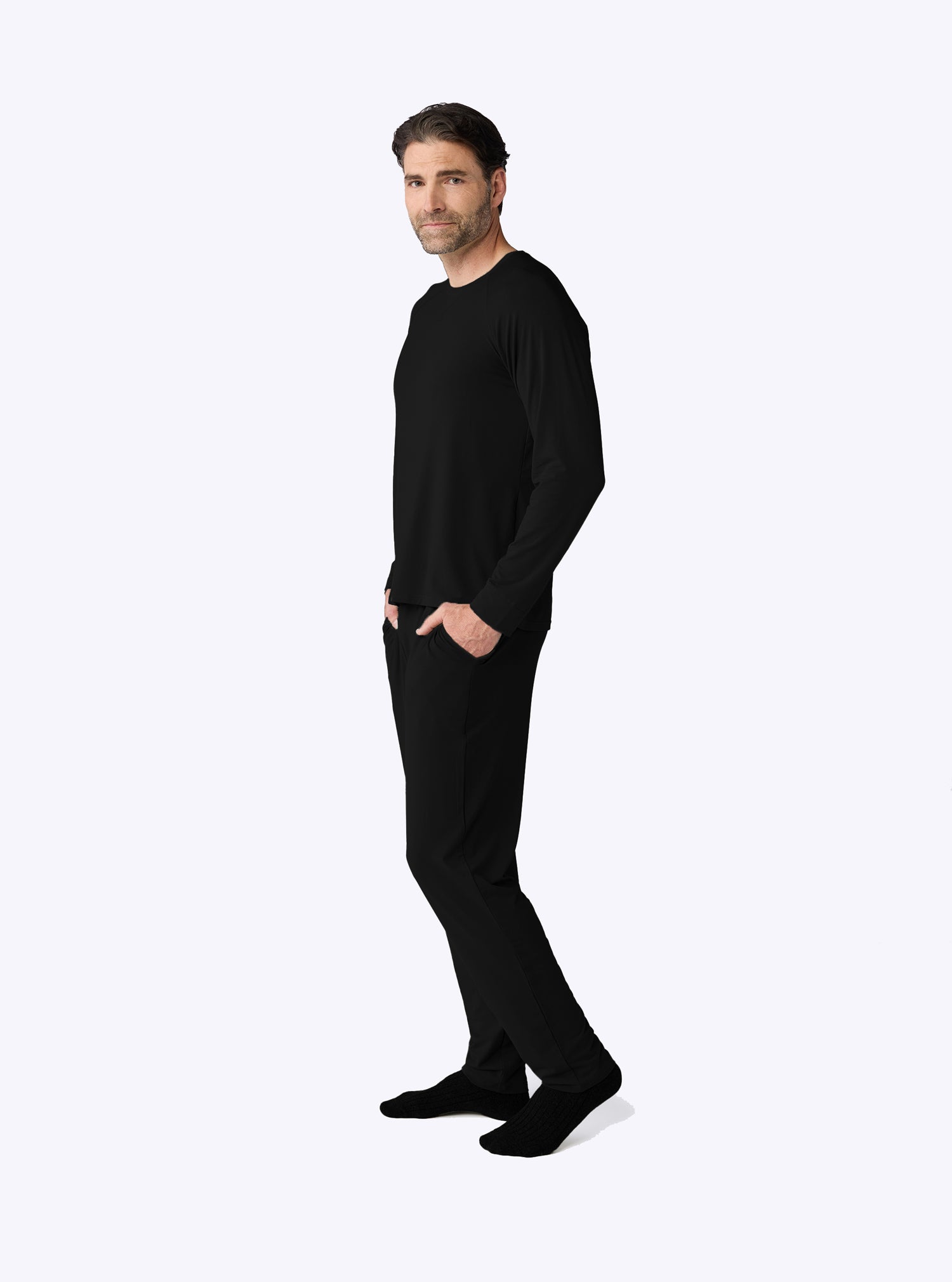 A man stands sideways with hands in pockets against a plain white background, wearing Cozy Earth's Men's Bamboo Stretch-Knit Long Sleeve Pajama Set in black. |Color:Black