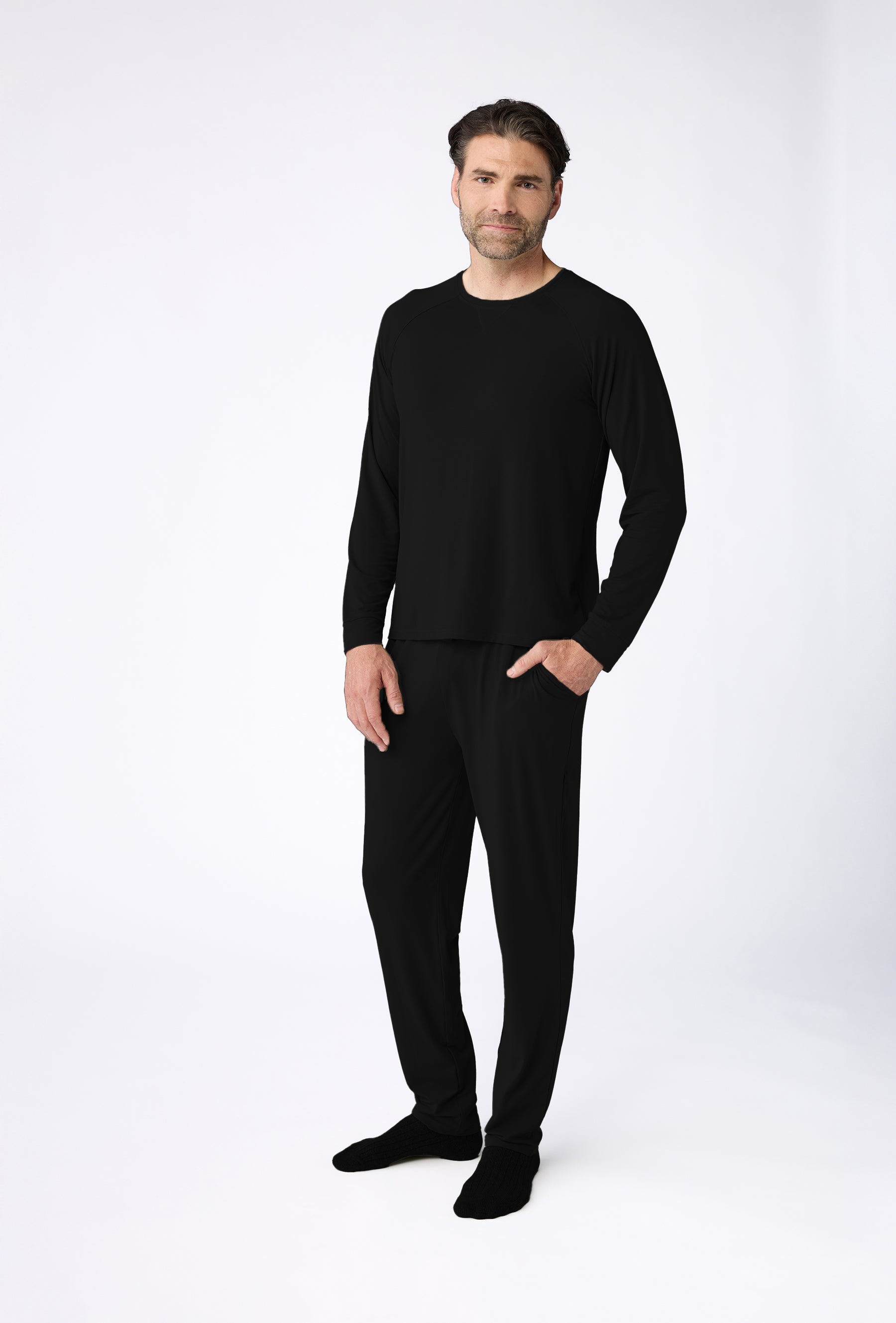 A man stands against a plain white backdrop, wearing the Cozy Earth Men's Bamboo Stretch-Knit Long Sleeve Pajama Set. He has short hair and a beard and gazes at the camera with a neutral expression, hands in pockets. |Color:Black