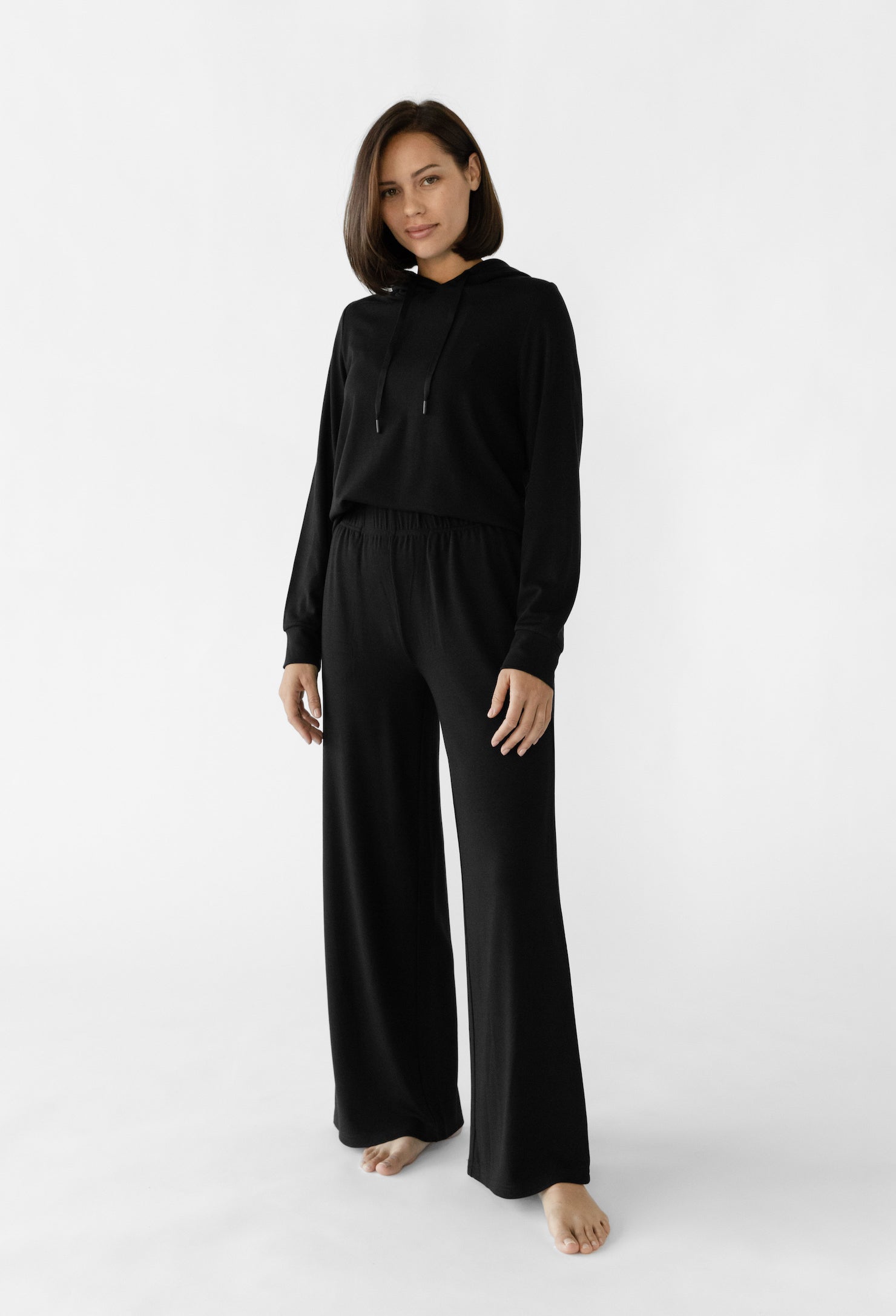 A person stands barefoot against a white backdrop, wearing Cozy Earth's Women's Ultra-Soft Bamboo Wide Leg Pull On Pant & Hoodie Set. They have short, dark hair and maintain a neutral expression with their hands at their sides. |Color:Black