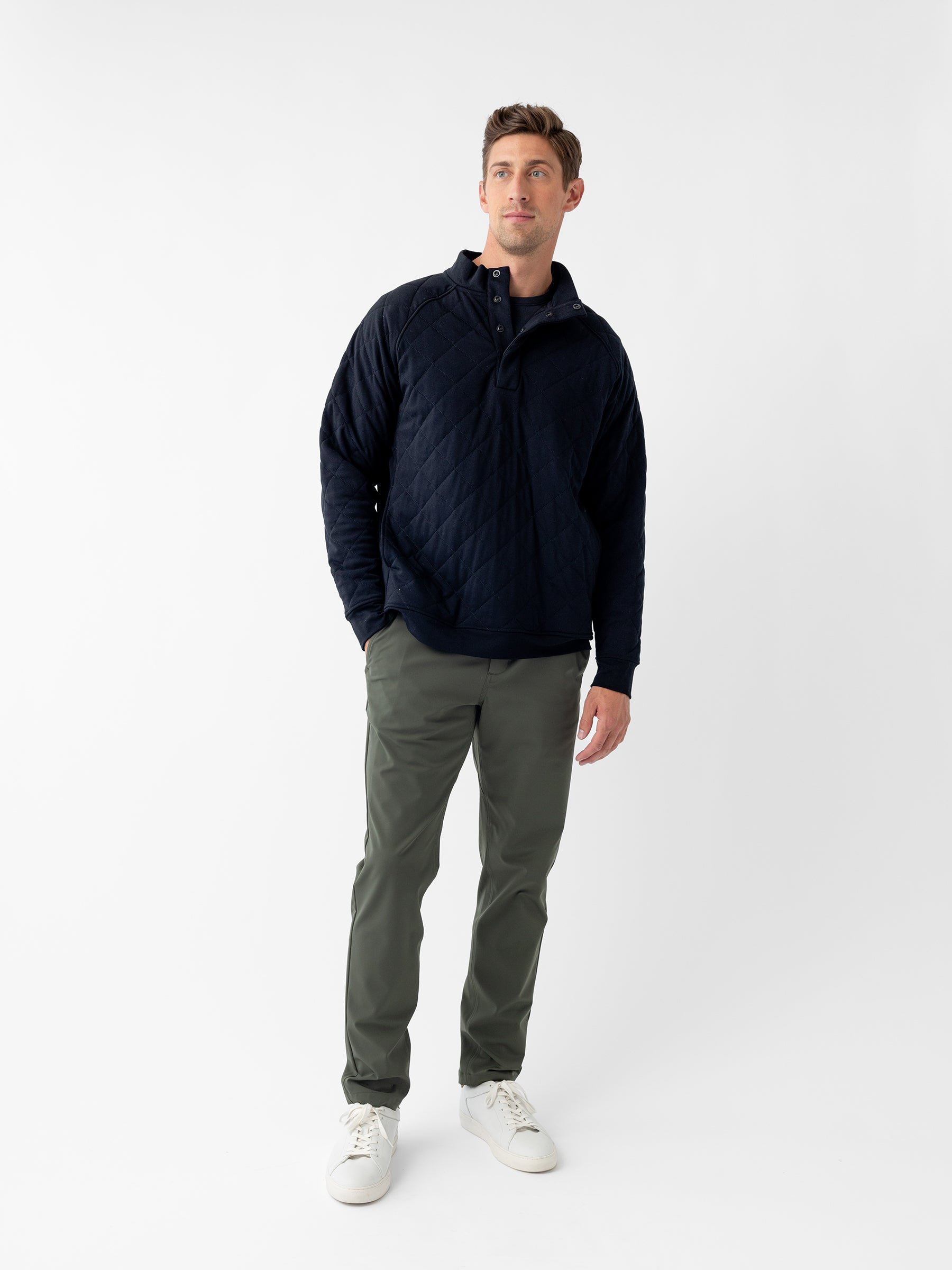 A man stands against a plain white background, wearing the Men's Ultra-Soft Bamboo Quilted Snap Pullover by Cozy Earth in navy blue, paired with olive green pants and white sneakers. He looks slightly to his right with a neutral expression, with his left hand by his side and his right hand in his pocket. |Color:Black