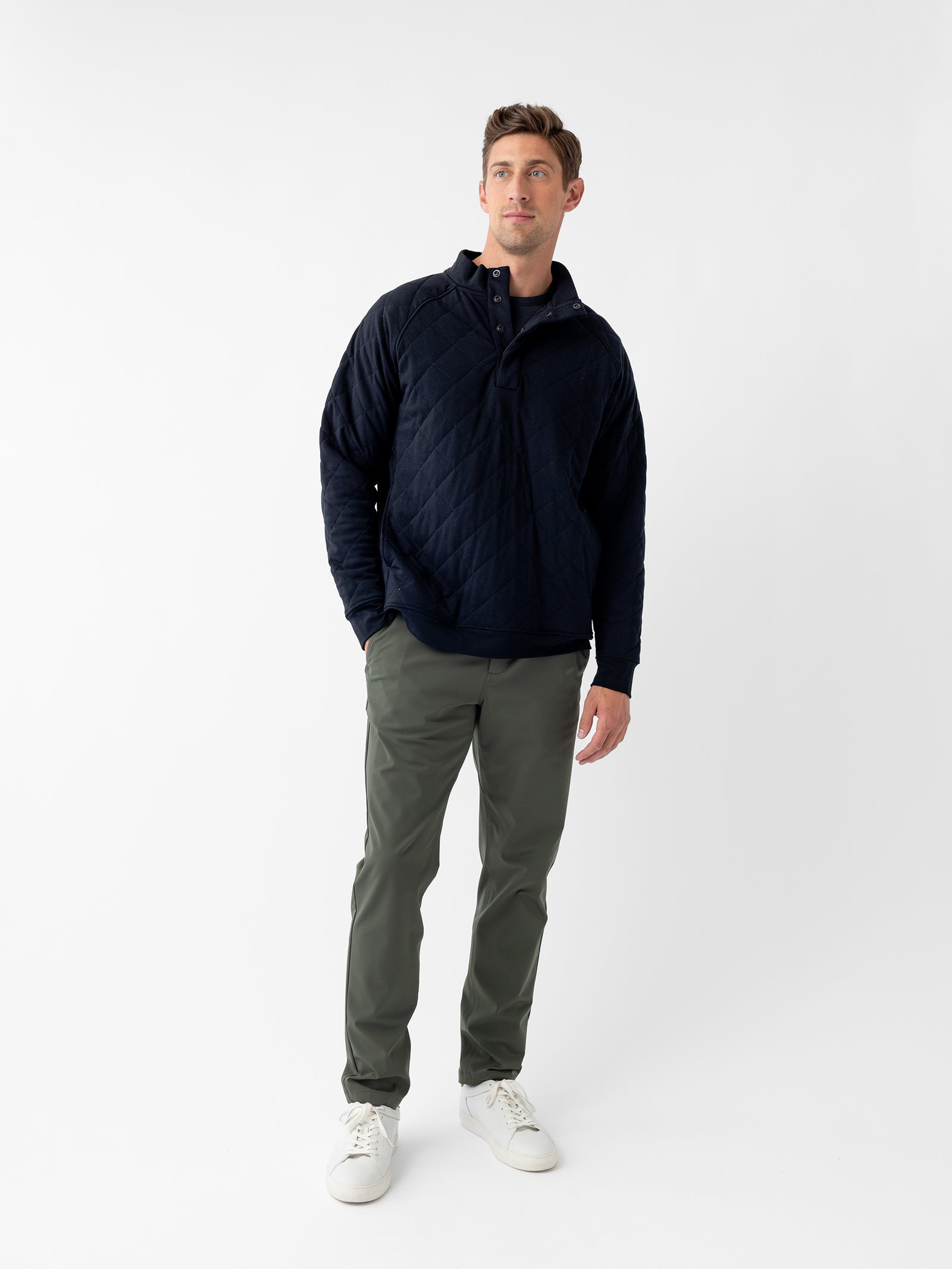 A man stands against a plain white background, wearing the Men's Ultra-Soft Bamboo Quilted Snap Pullover by Cozy Earth in navy blue, paired with olive green pants and white sneakers. He looks slightly to his right with a neutral expression, with his left hand by his side and his right hand in his pocket. 