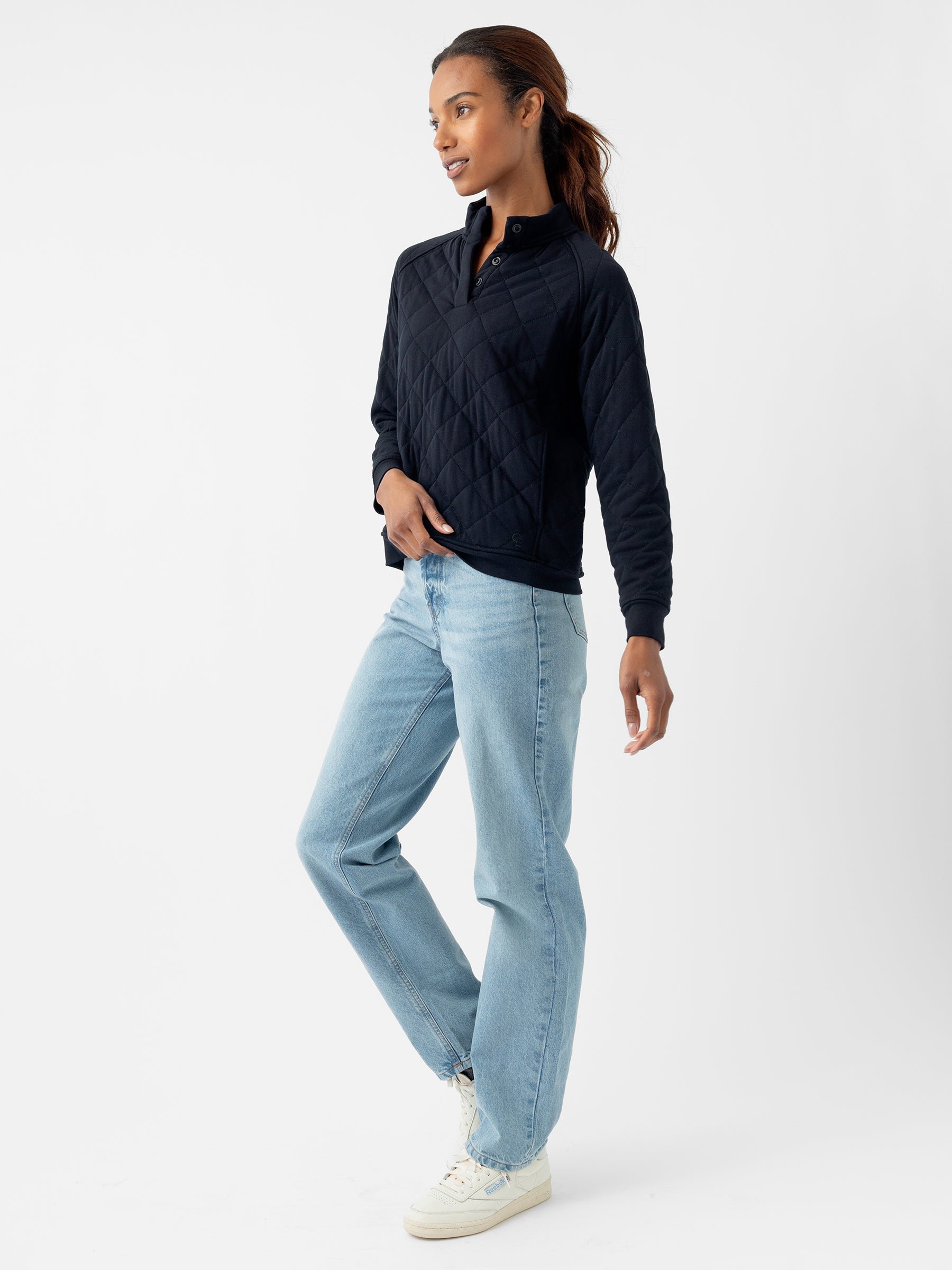 A person with long dark hair is wearing the Cozy Earth Women's Ultra-Soft Bamboo Quilted Snap Pullover in black and light blue jeans. They are standing casually with one foot slightly forward and one hand resting on their hip against a white background. 