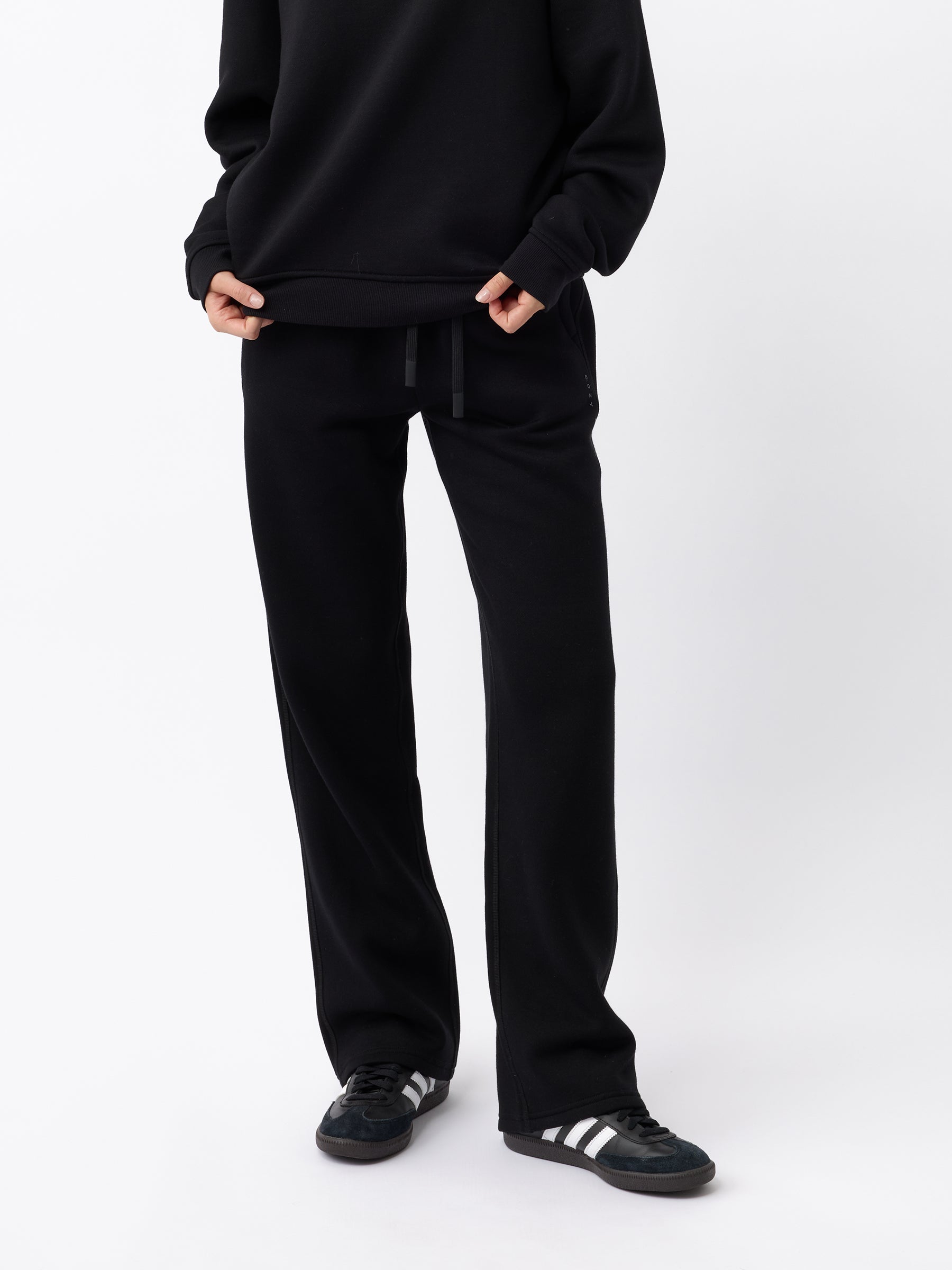 A person is standing against a plain white background, wearing Cozy Earth's Women's CityScape Wide Leg Pant in black and a matching black sweater. They are holding the bottom of their sweater with both hands. Their feet are adorned with black sneakers featuring white stripes. |Color:Black