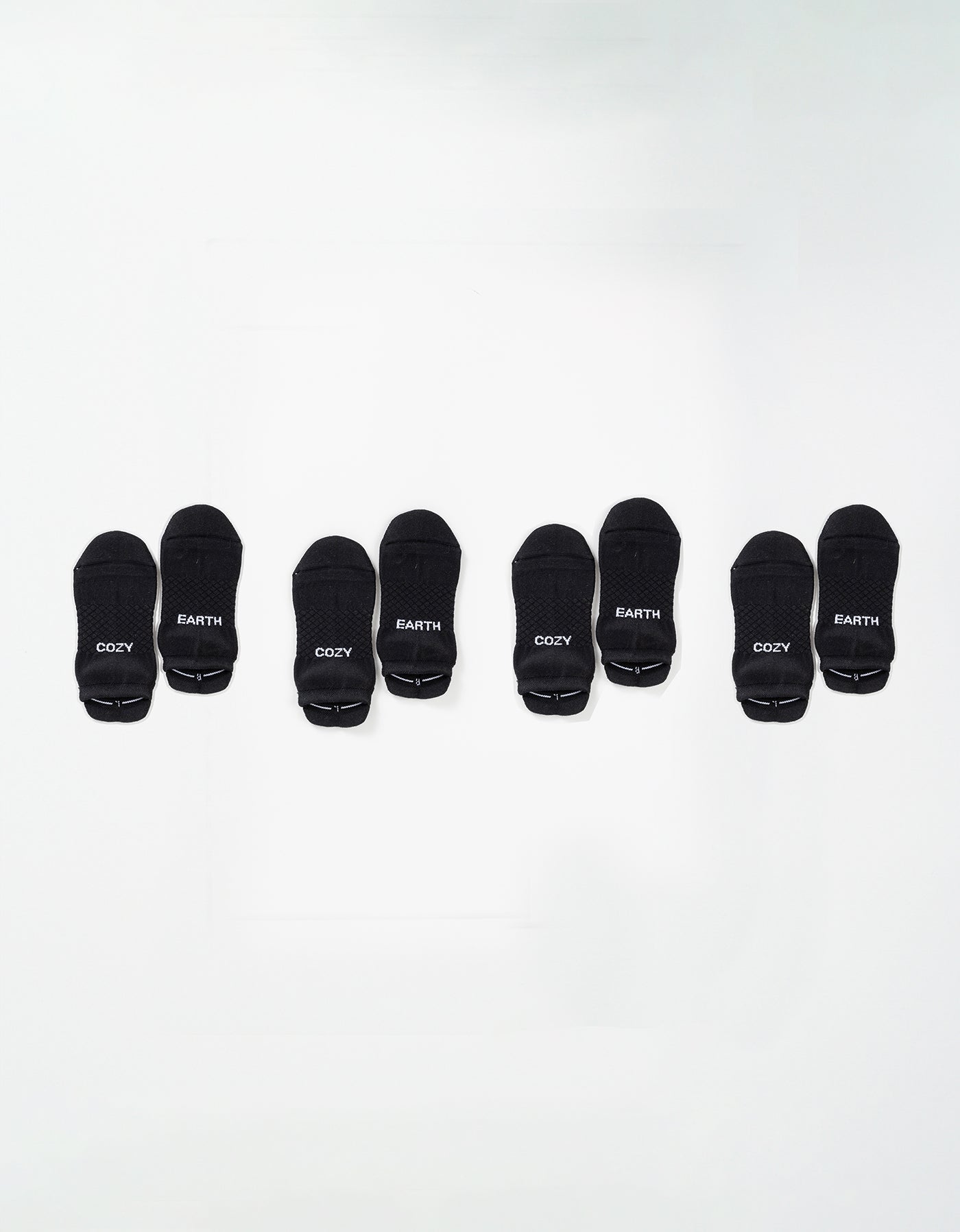 Four pairs of black Essential Ankle Socks by Cozy Earth are arranged in a row on a white background, each pair featuring "COZY" and "EARTH" in white text on the toes. 