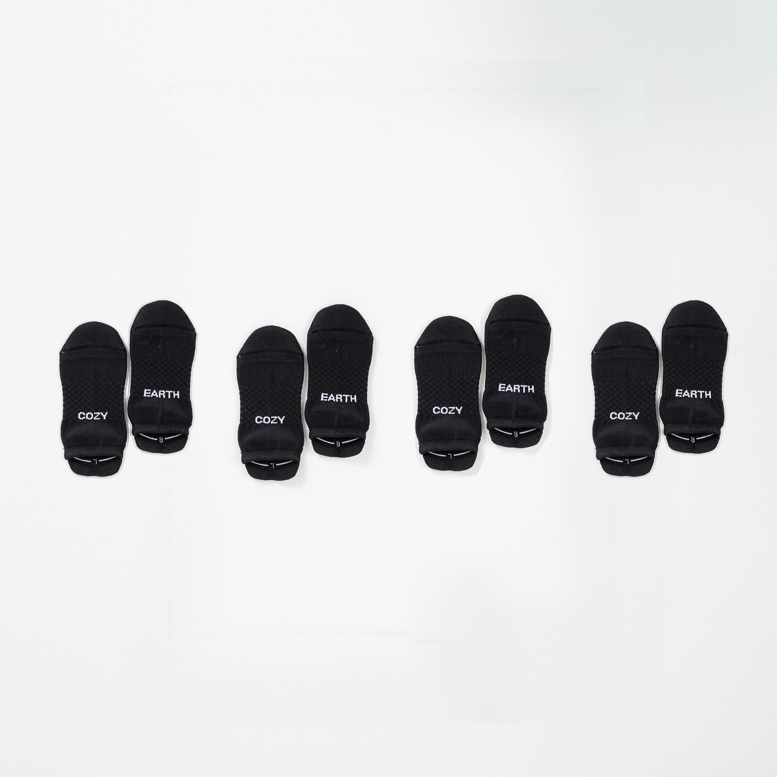 A row of socks from the Essential Ankle Sock 4-Pack by Cozy Earth is arranged on a white surface. The black ankle socks are lined up in pairs, each featuring the words "COZY" or "EARTH" written in white text. There are 8 socks in total, forming 4 pairs. 