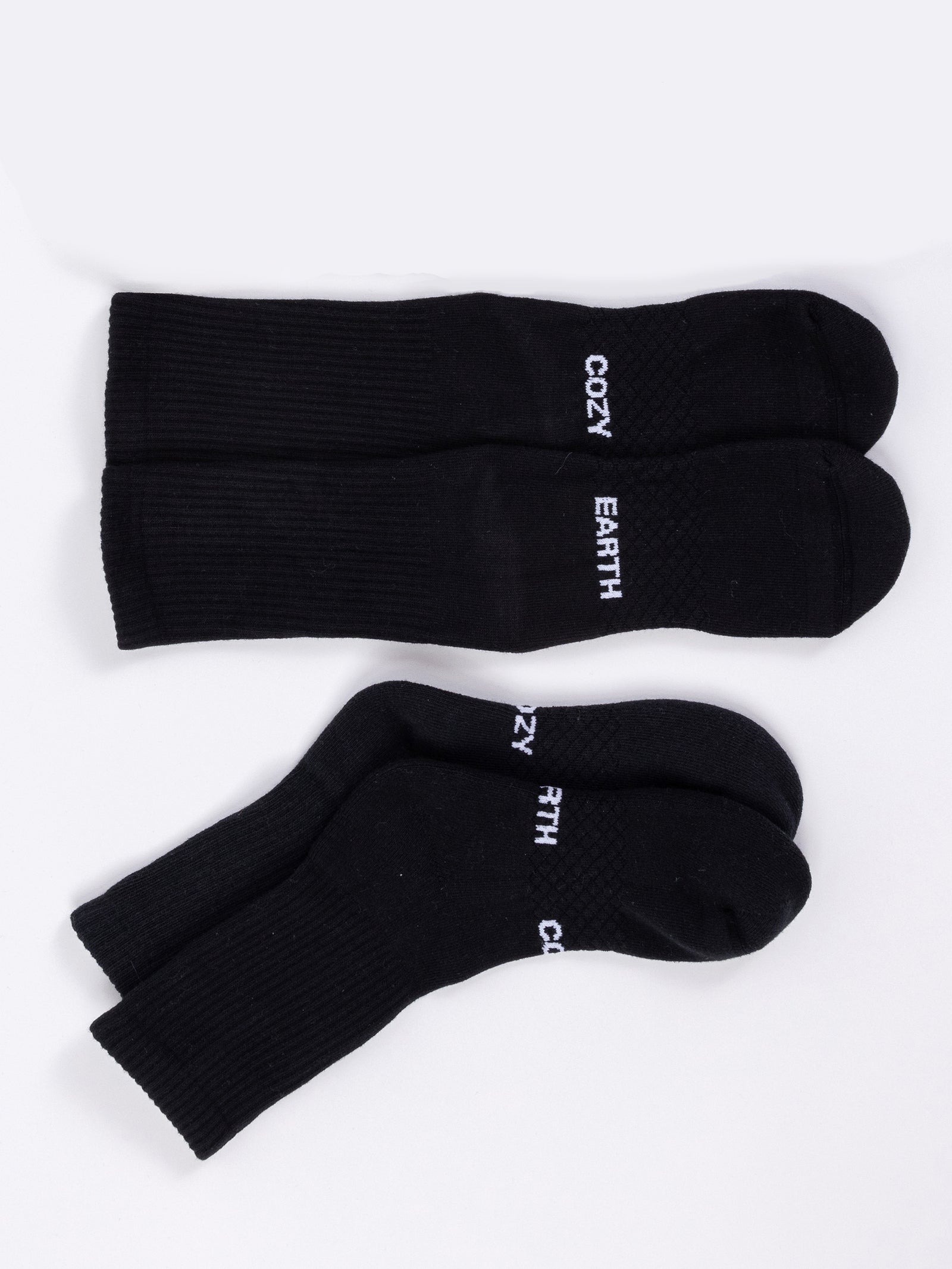 A pair of Essential Calf Socks from the Cozy Earth brand is displayed on a white surface. One sock is laid flat, revealing the top side with "COZY EARTH" printed in white, while the other sock is stacked, draped over its pair, also showcasing the "COZY EARTH" text. 