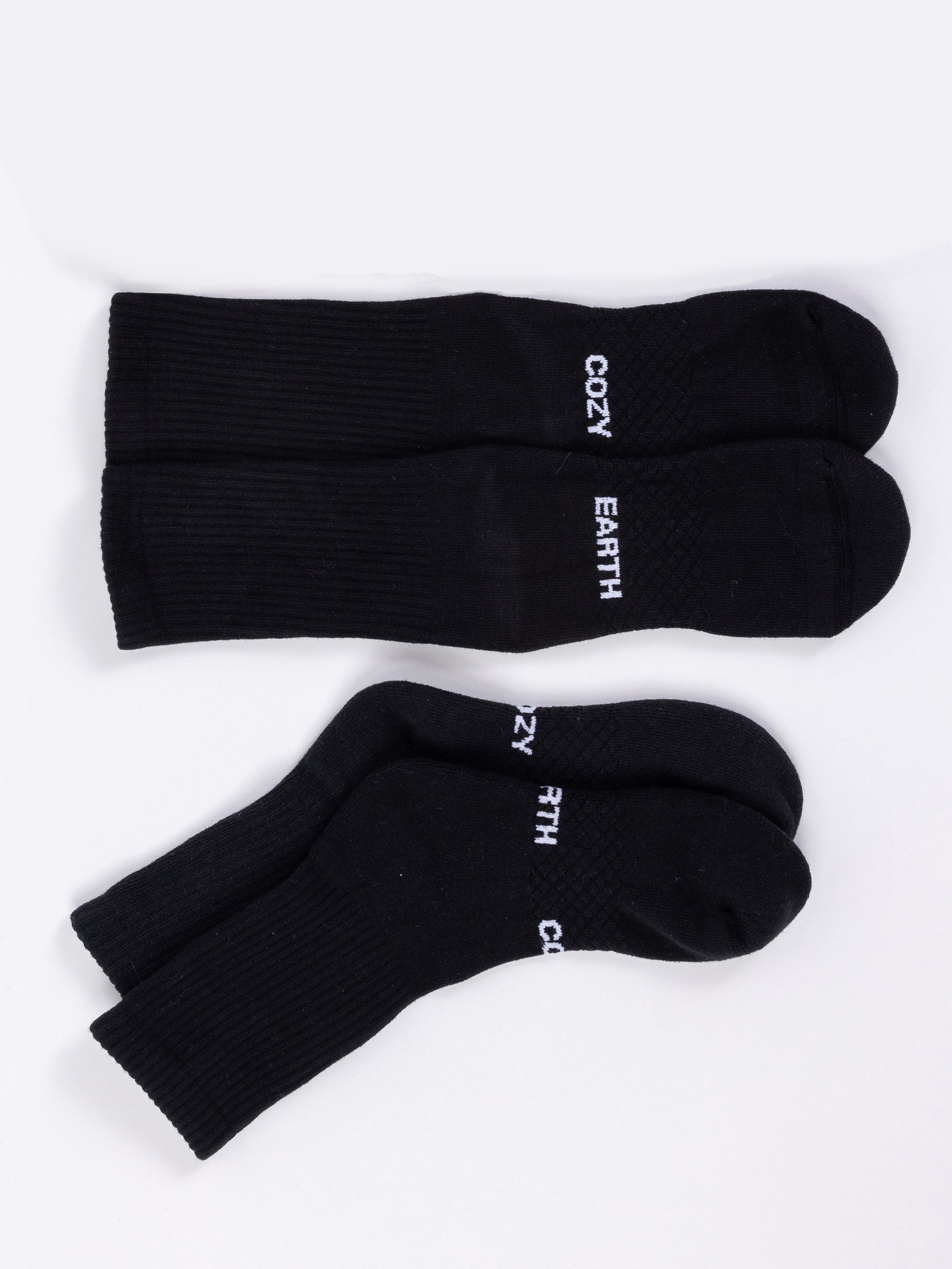 A pair of Essential Calf Socks from the Cozy Earth brand is displayed on a white surface. One sock is laid flat, revealing the top side with "COZY EARTH" printed in white, while the other sock is stacked, draped over its pair, also showcasing the "COZY EARTH" text. |Color:Black