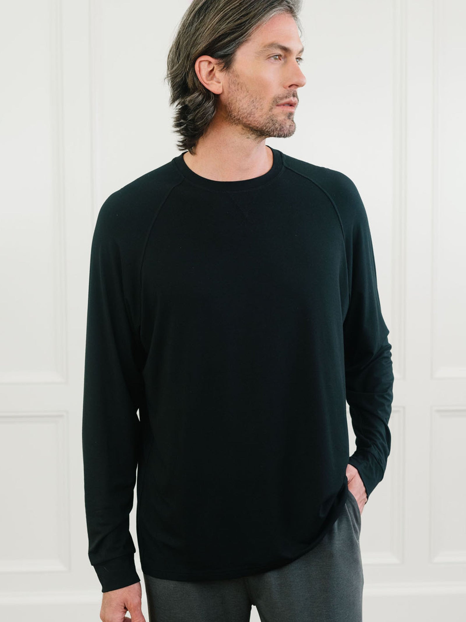 A man with shoulder-length gray hair and a beard stands against a white paneled wall. He is wearing a Cozy Earth Men's Stretch-Knit Long Sleeve Bamboo Pajama Jogger Set in black, with his right hand in his pocket. He is gazing off to the side.  