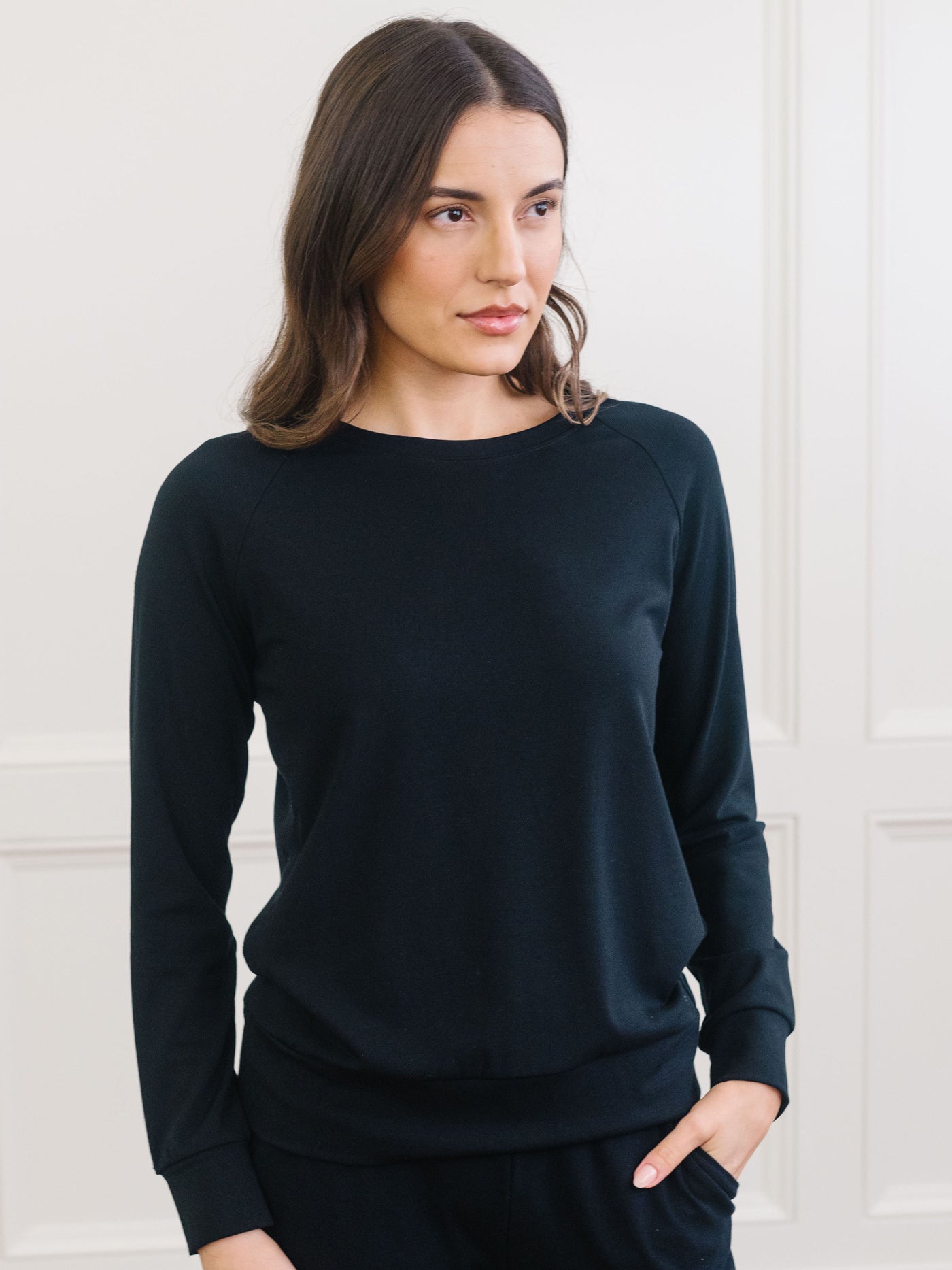 A woman with long brown hair wears the black pullover from Cozy Earth's Women's Ultra-Soft Bamboo Wide Leg Pull On Pant & Pullover Crew Set against a light-colored wall, slightly looking right with her hands in her pockets. 