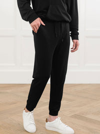 A person wearing Cozy Earth's Men's Brushed Bamboo Jogger Pant in black stands on a light wood floor before a white paneled wall. Their face is not visible.