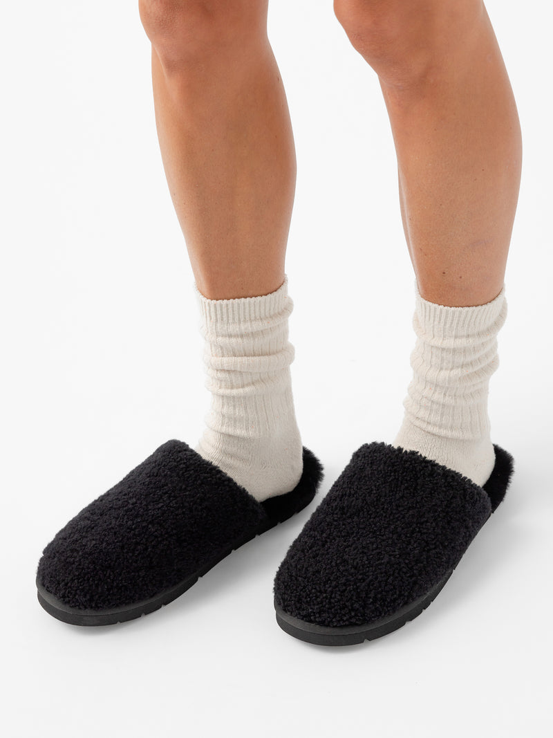 Close-up of a person's lower legs wearing white crew socks and Cozy Earth's Puffy Sheep Slipper, which are black and fuzzy. The background is plain white.