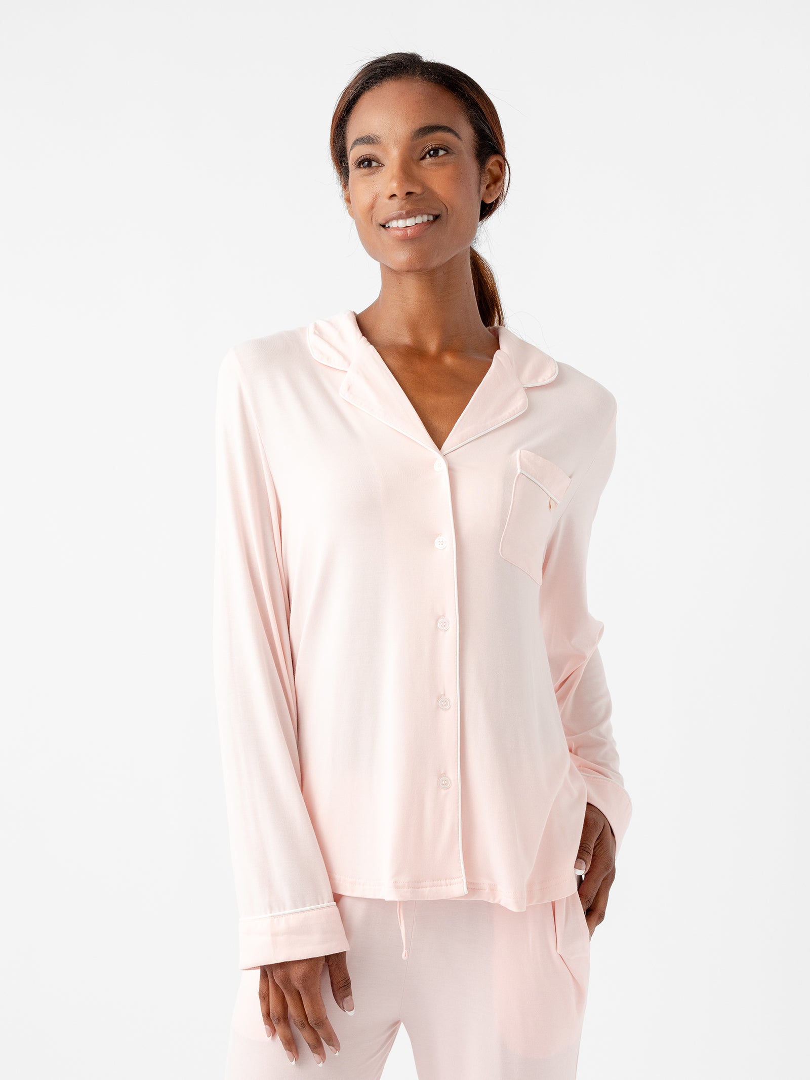 A woman is smiling and wearing a light pink Women's Long Sleeve Bamboo Pajama Top in Stretch-Knit TALL from HIDE, complemented by matching pants. The top features a button-up design with a single chest pocket adorned with subtle white trim. The background is plain white. 