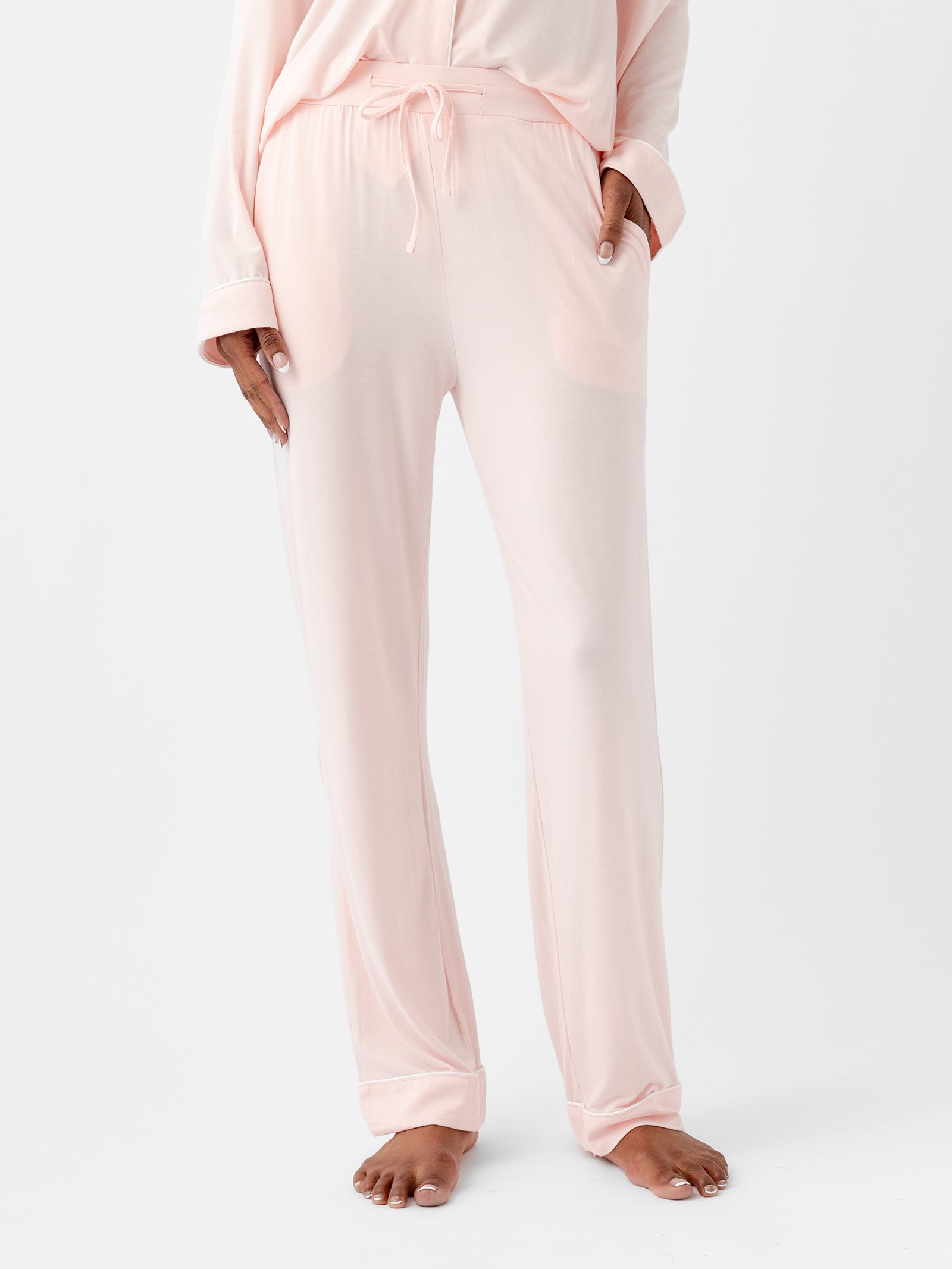 A woman wearing HIDE Women's Stretch-Knit Bamboo Pajama Pants in light pink stands with one hand in her pocket. The loose-fitting pants feature an elastic waistband and a drawstring tie. Her bare feet are visible against a plain white background. 