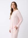 A woman with long, wavy hair stands against a plain white background, wearing a light pink Women's Stretch Knit Bamboo Long Sleeve Lounge Tee and matching pants by Cozy Earth, with one hand in the pocket of the pants. |Color:Blossom