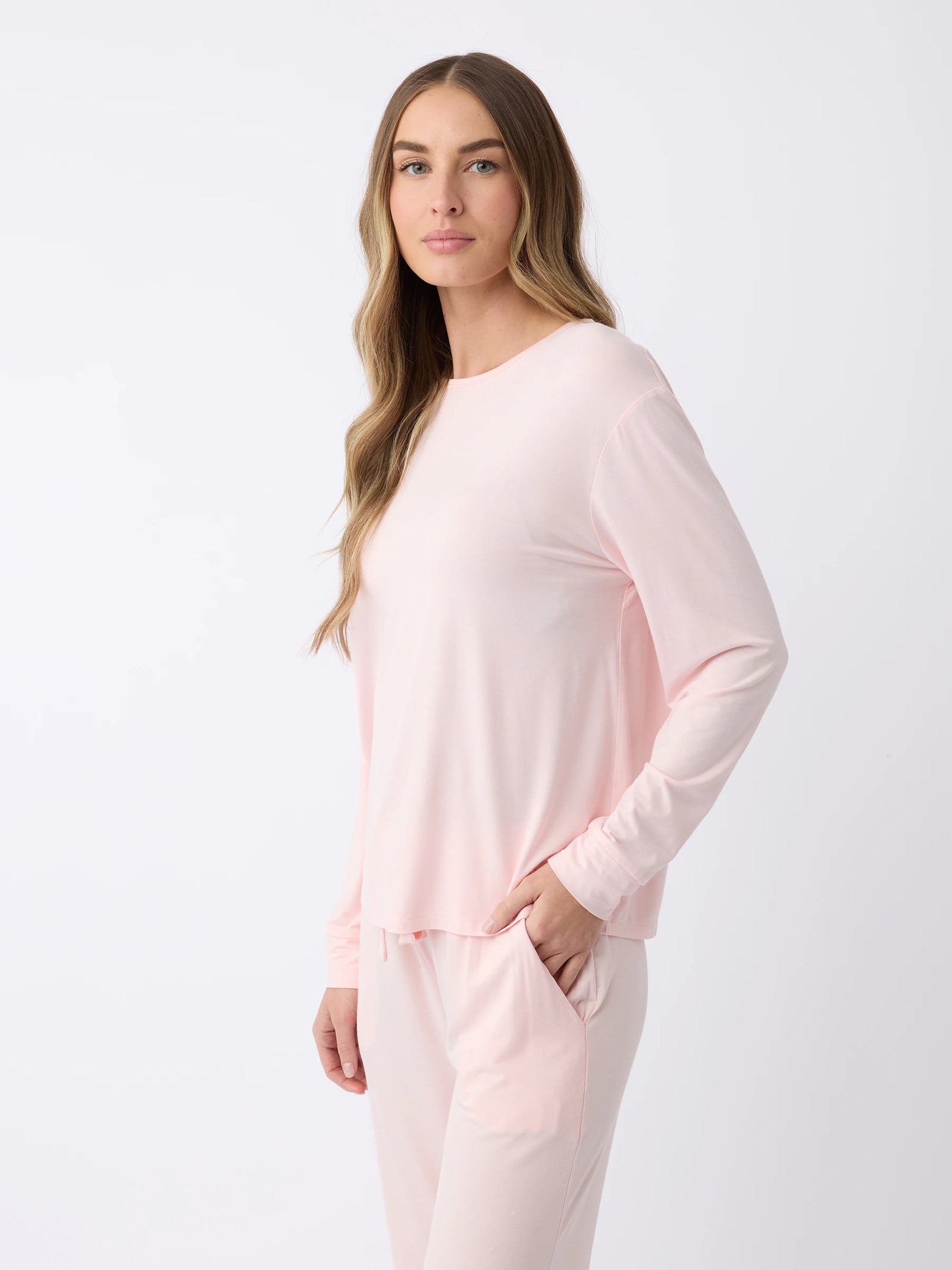 A woman with long, wavy hair stands against a plain white background, wearing a light pink Women's Stretch Knit Bamboo Long Sleeve Lounge Tee and matching pants by Cozy Earth, with one hand in the pocket of the pants. 