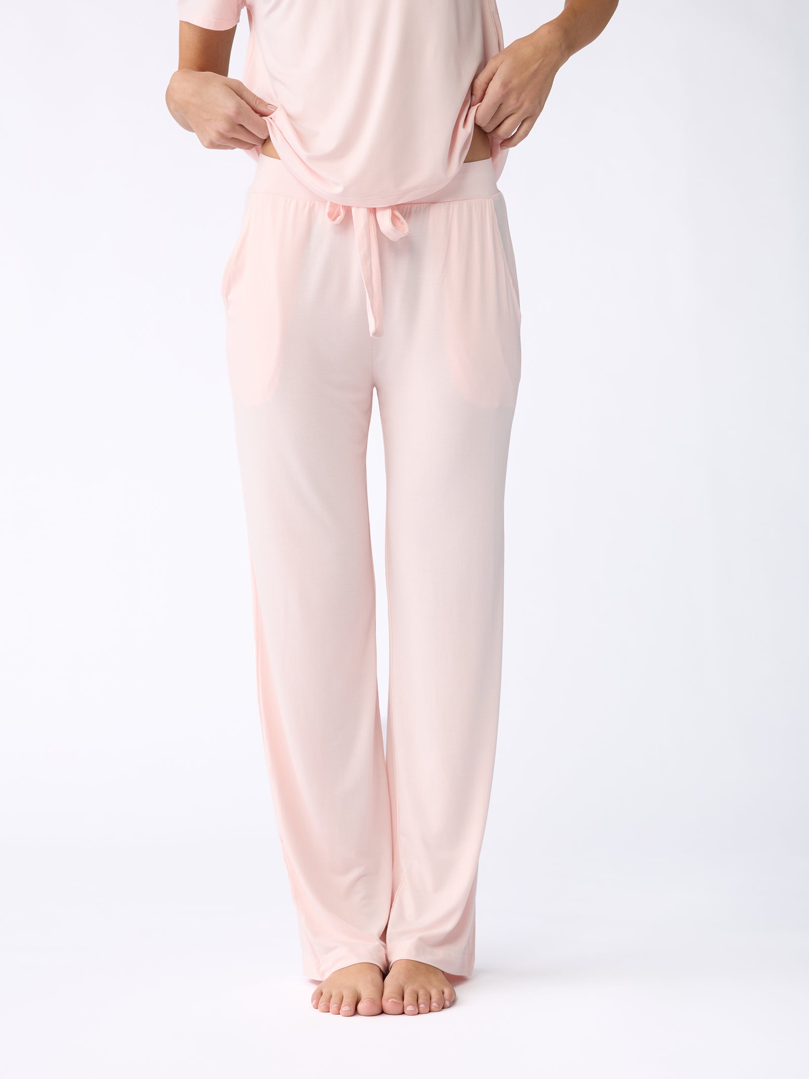 A person is seen barefoot against a plain white background, wearing a soft and comfortable Cozy Earth ensemble featuring the Stretch Knit Bamboo Pant in light pink with a long-sleeved top to match. The individual's face is not visible. 