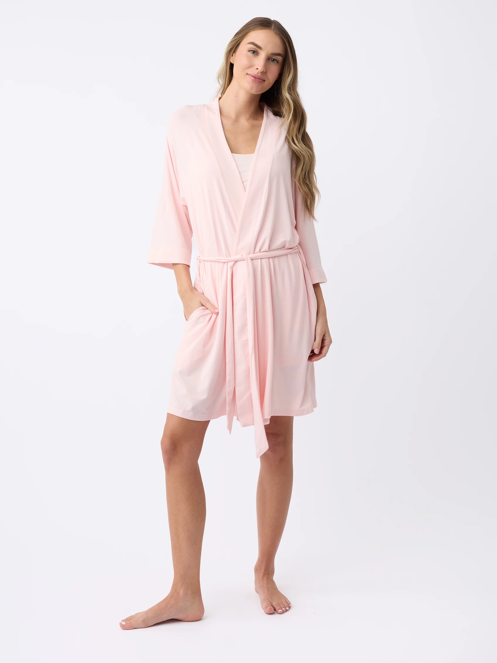 A woman with long hair stands barefoot against a plain white background, wearing the Women's Stretch-Knit Bamboo Kimono Robe by Cozy Earth. The light pink robe features three-quarter sleeves and a tied waist. She poses casually with a slight smile. |Color:Blossom