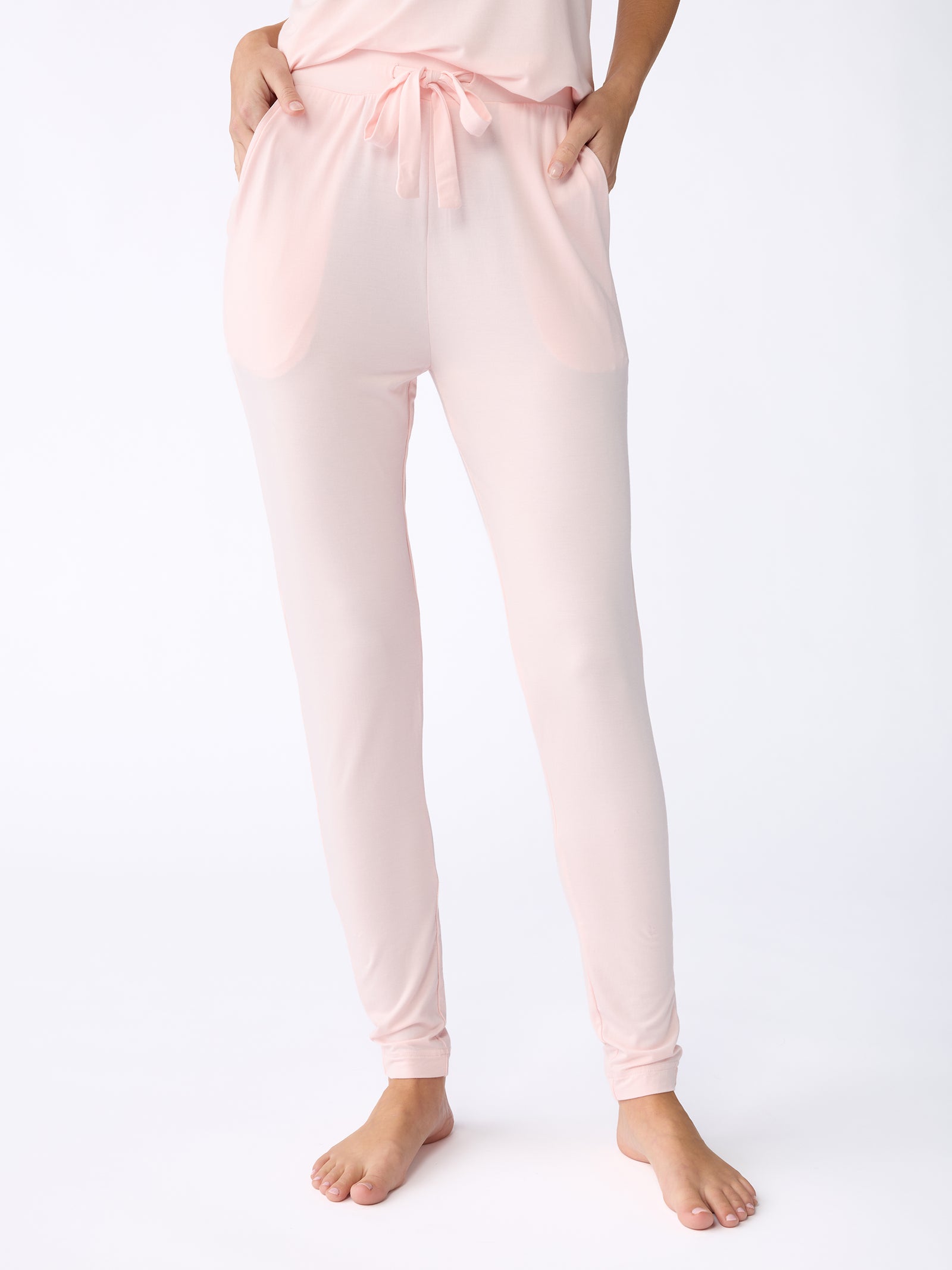 A person is standing barefoot wearing Cozy Earth's Stretch Knit Bamboo Tapered Pant in light pink, featuring a drawstring waist and two pockets. The background is plain white. 