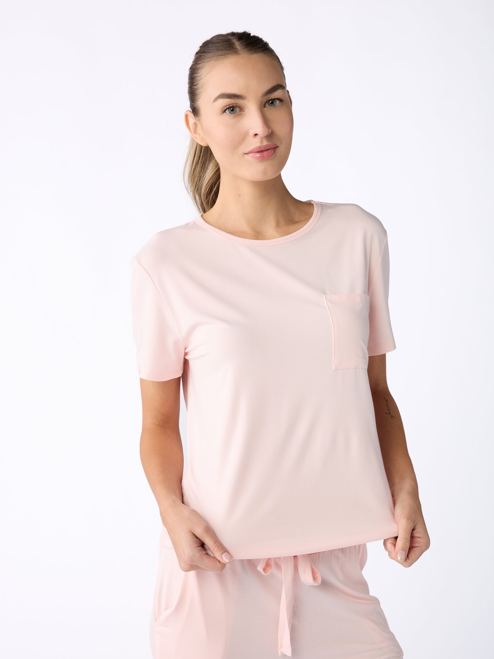 A person dressed in a Women's Stretch-Knit Bamboo Lounge Tee from Cozy Earth, in light pink, and matching pants stands against a plain white background. The tee features a pocket on the left side. The person looks forward with a slight smile, gently holding the bottom of the tee. 